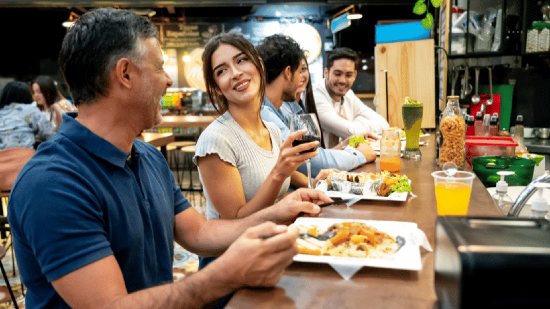 Personas disfrutando de una comida en un mostrador en un ambiente de comedor informal. Una mujer está sonriendo y sosteniendo una copa de vino mientras conversa con un hombre a su lado. Platos de comida y bebidas están dispuestos frente a ellos.