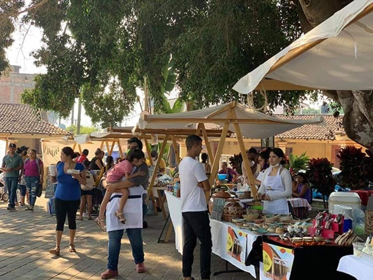 Personas comprando en un mercado al aire libre, con vendedores ofreciendo diversos productos bajo toldos sombreados.
