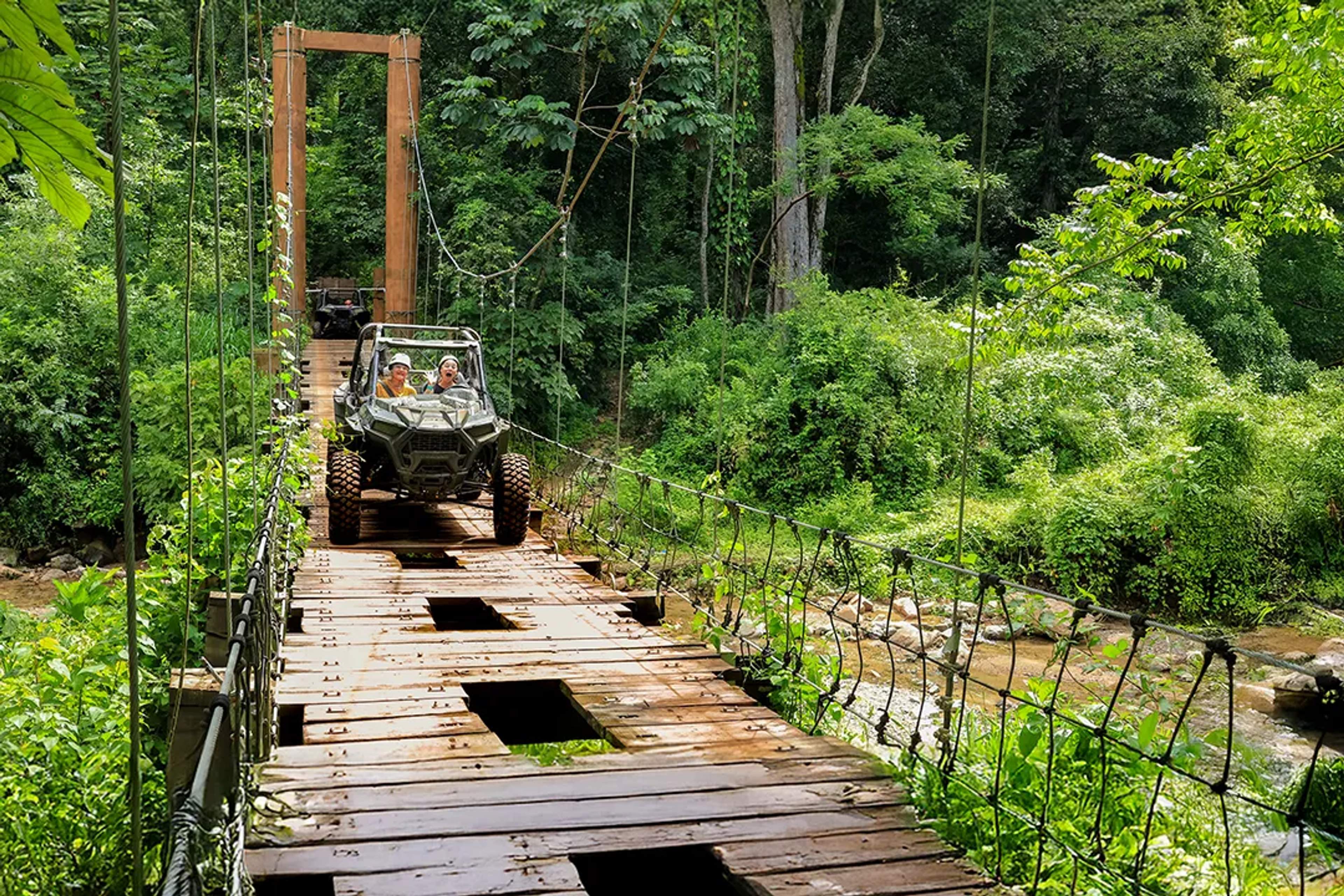 Two people cross a wooden suspension bridge in a Polaris® RZR off-road vehicle during a jungle adventure, surrounded by lush greenery and a stream below.
