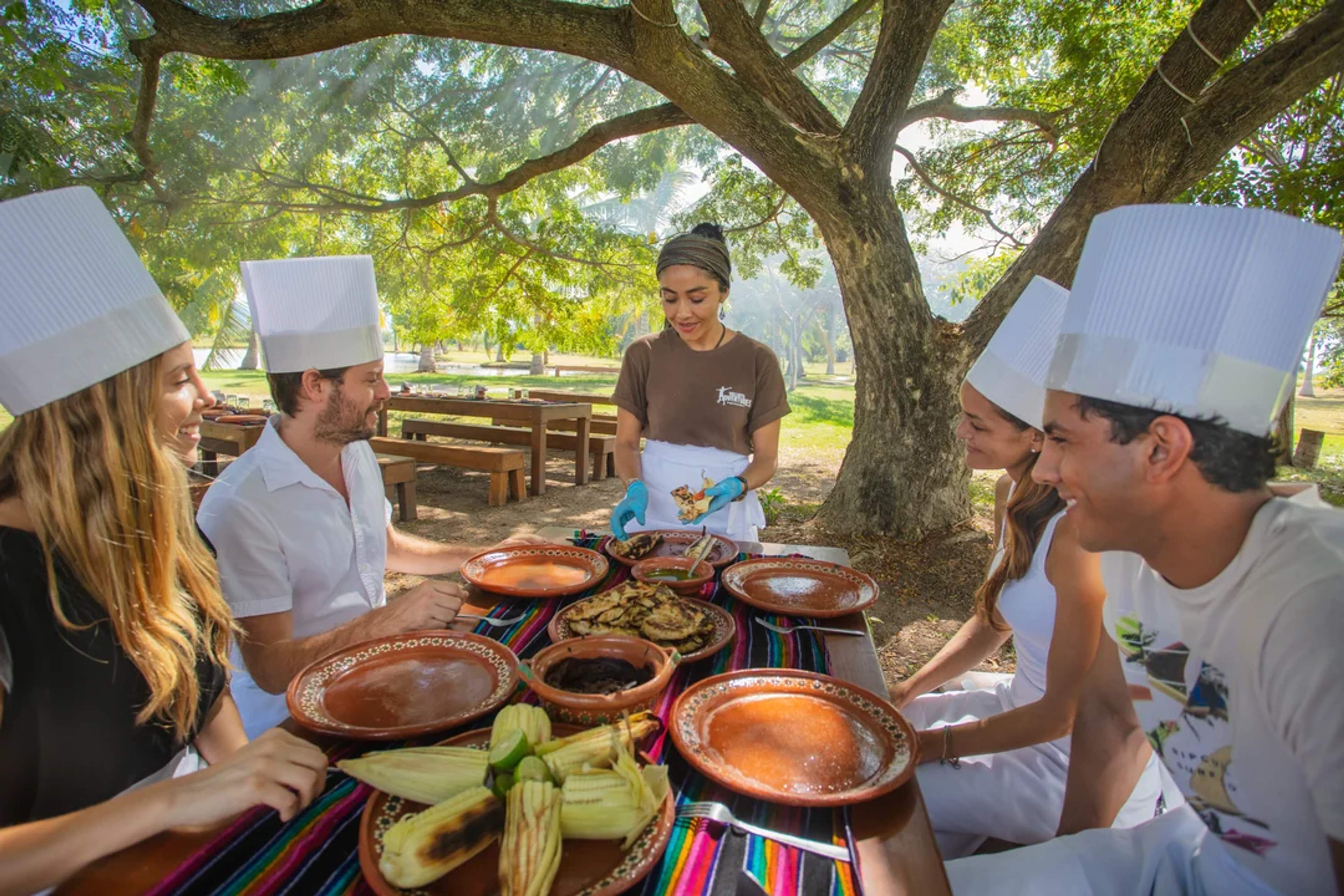 A group enjoys an outdoor cooking class under a tree, wearing chef hats and learning traditional recipes together.