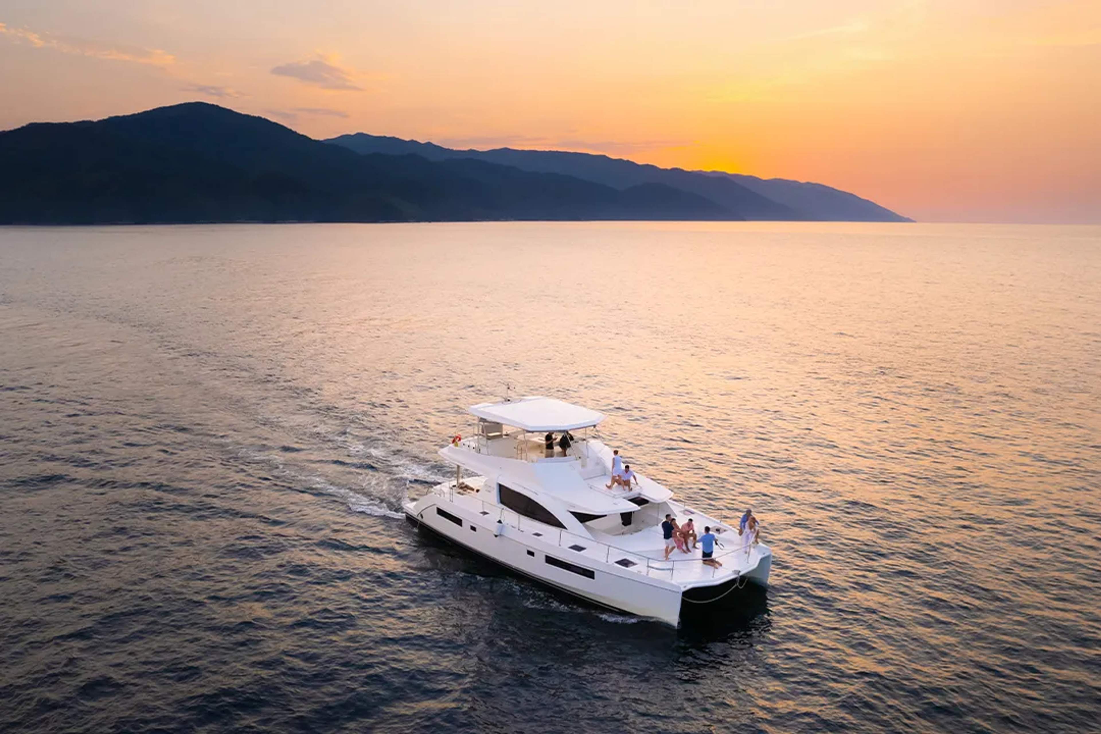 Luxury yacht sailing at sunset with friends aboard, calm sea and mountains in the background.