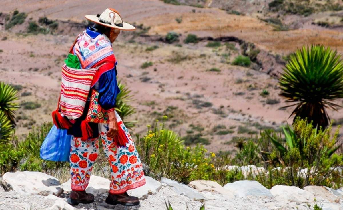 Huichol indigenous person in traditional colorful clothing walking in a rocky landscape.