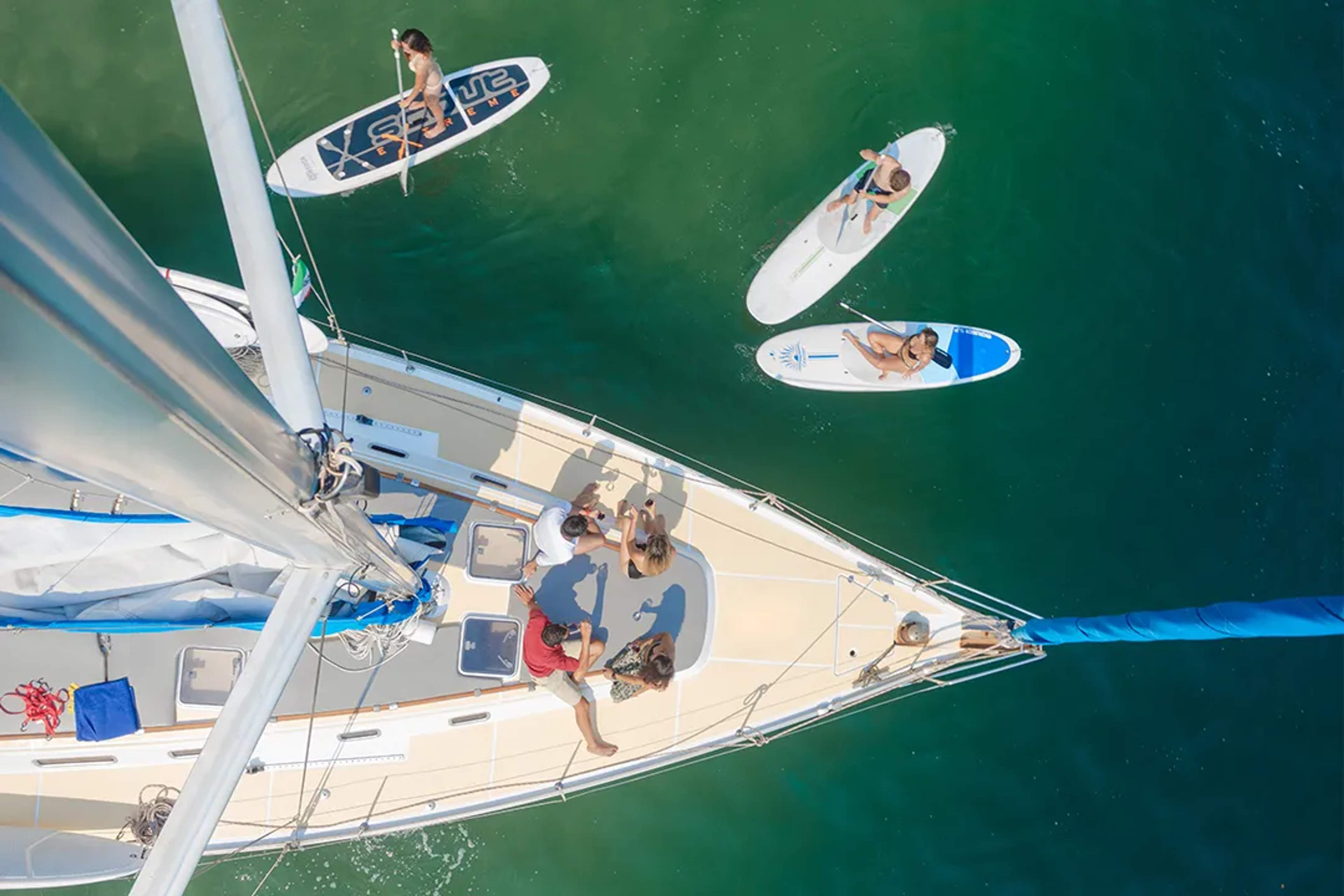 Aerial view of people relaxing on a sailboat while others paddleboard in clear green water.