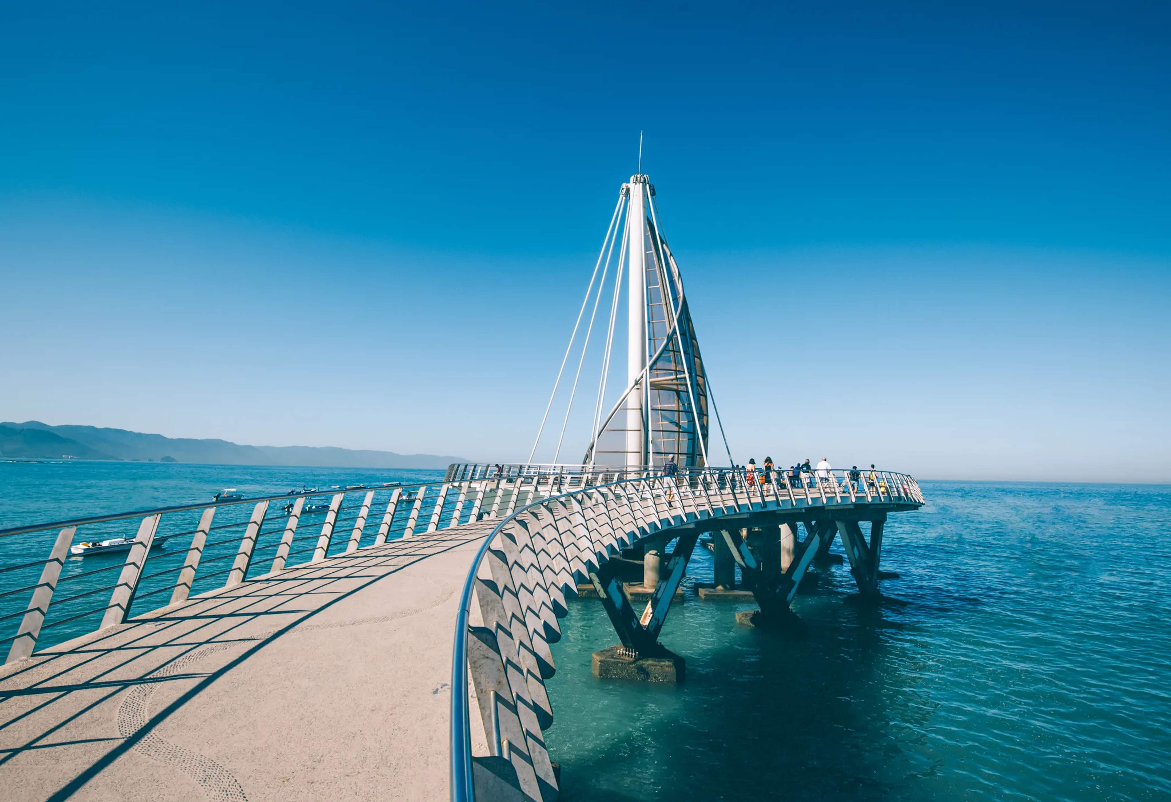 A modern curved pier with white railings stretching into clear turquoise water near Puerto Vallarta under a bright blue sky