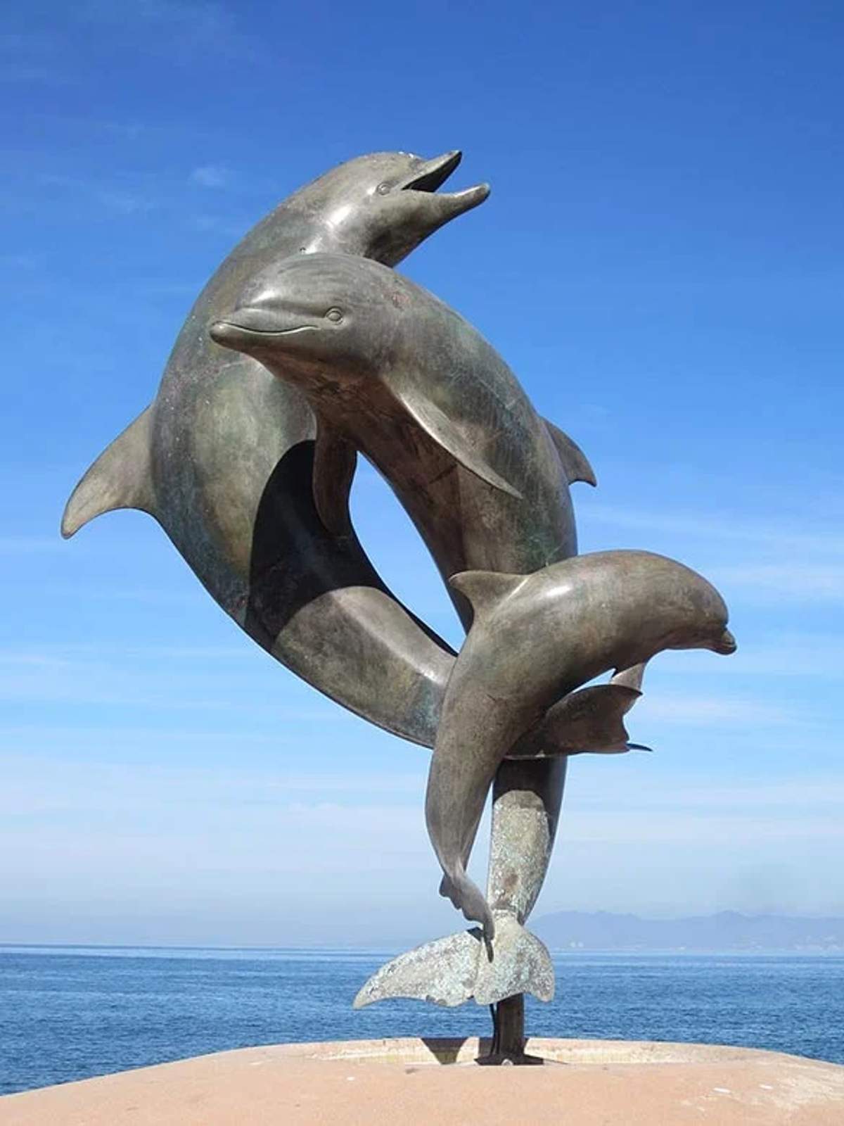 Escultura de tres delfines saltando juntos, con el cielo azul claro y el océano de fondo.