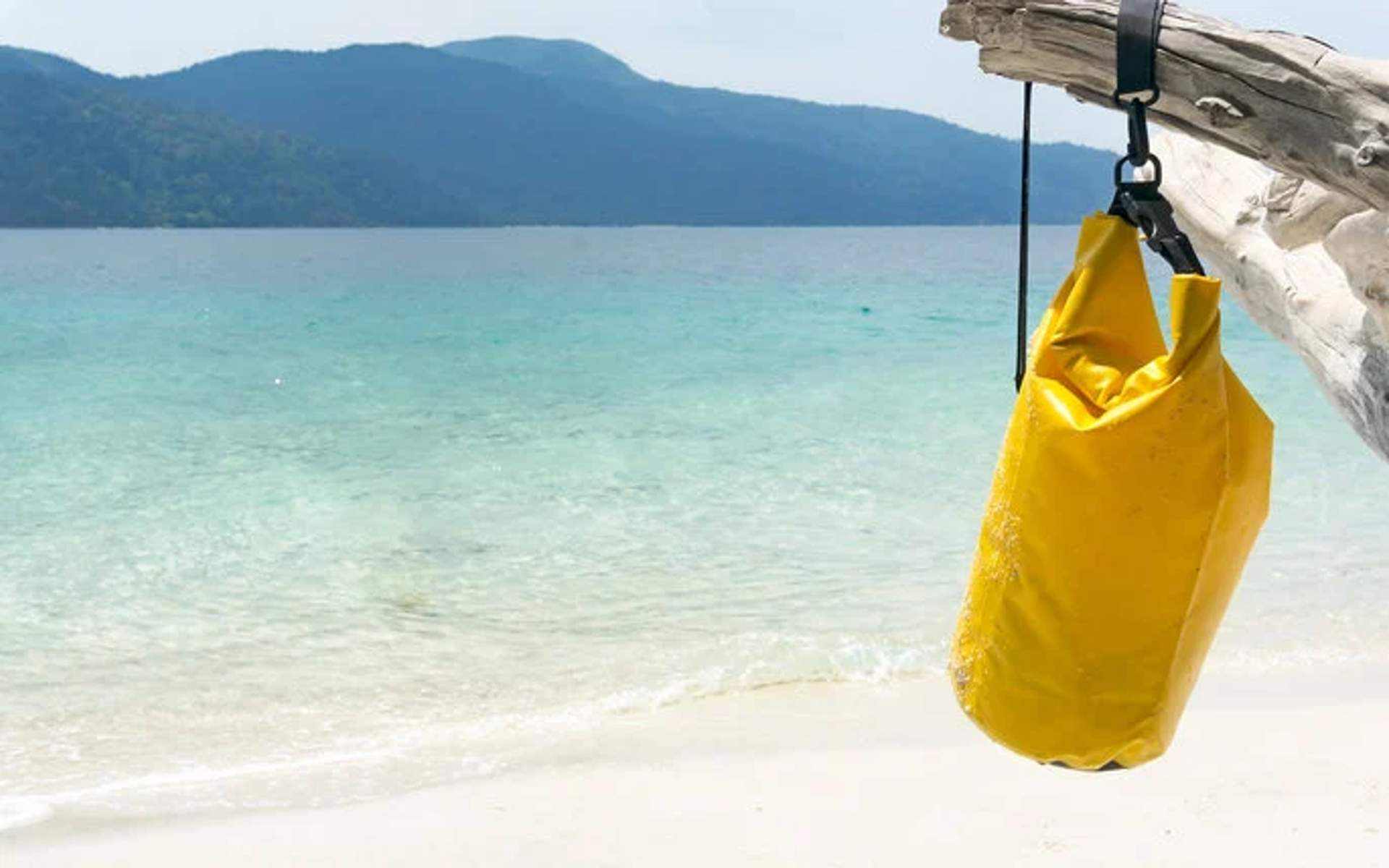 Yellow waterproof bag hanging from a tree branch on a beach with clear water and mountains in the background.