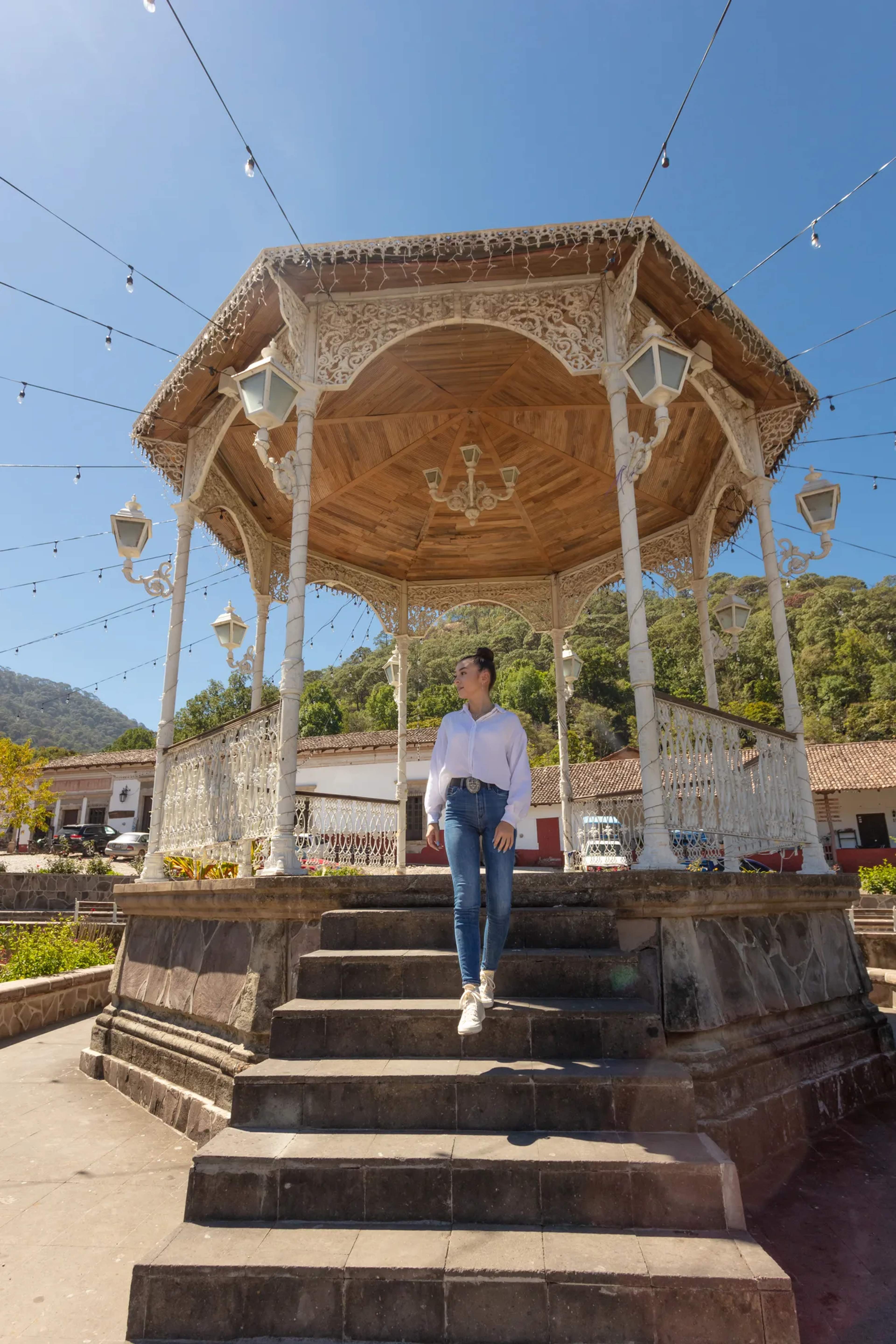 A visitor stands beneath an ornate wooden gazebo in San Sebastián's town plaza, surrounded by green hillsides and colonial