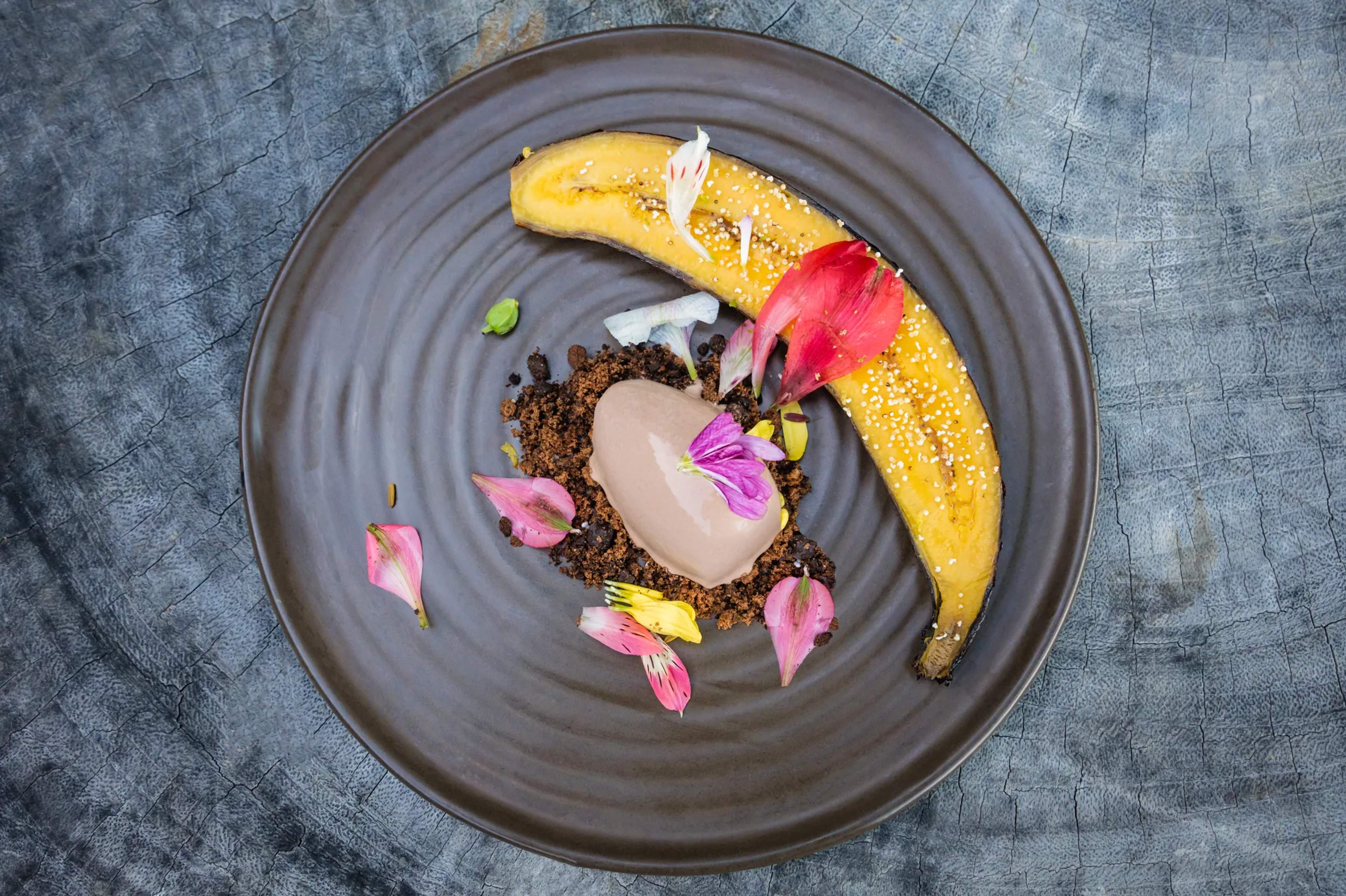 Gourmet dessert plate with caramelized banana, creamy mousse, chocolate crumble, and edible flowers on a dark ceramic dish.