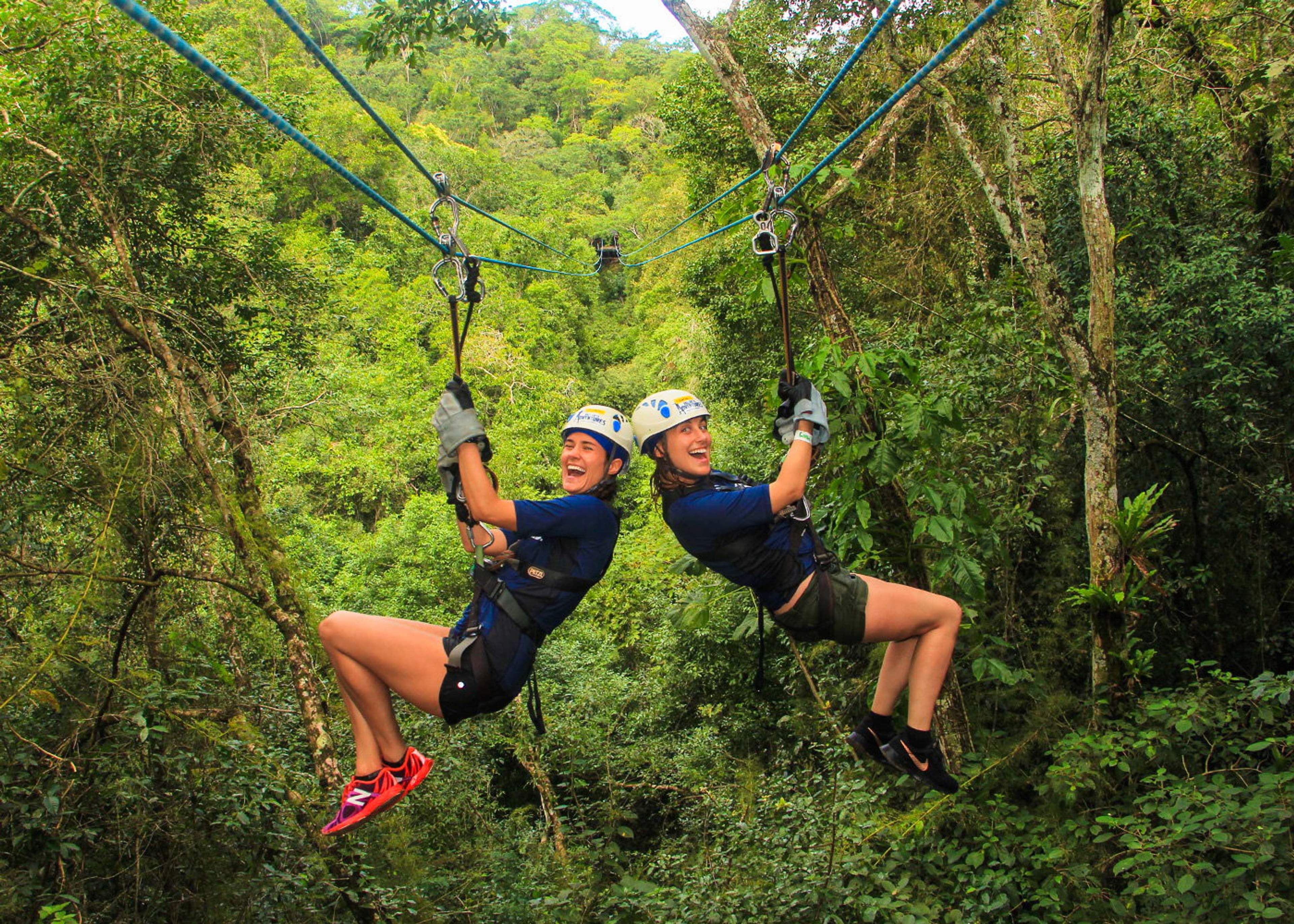 Two women wearing helmets and harnesses ziplining side by side through a lush forest, laughing and enjoying the adventure.