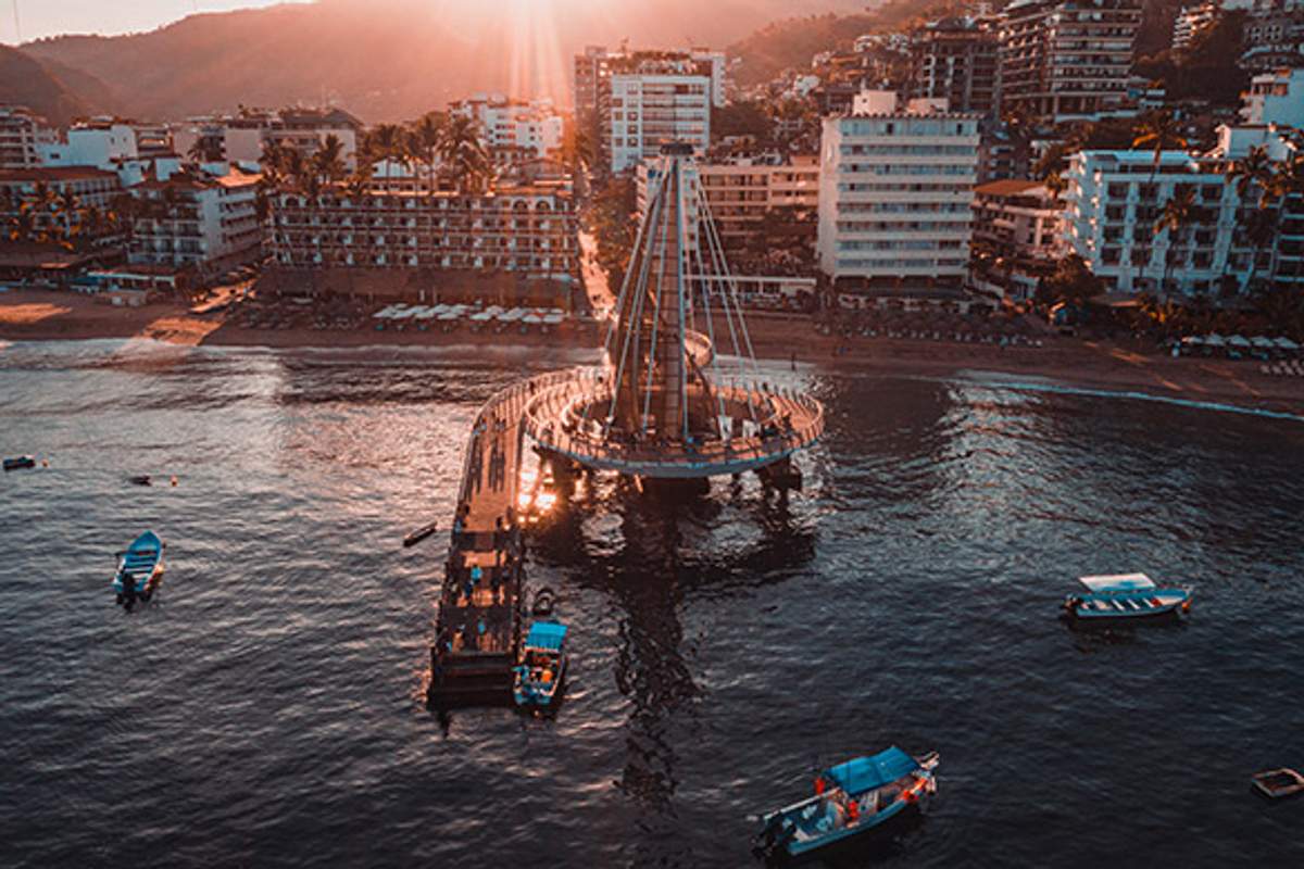 Vista al atardecer del Muelle de Los Muertos en Puerto Vallarta con botes en el agua y la ciudad al fondo.