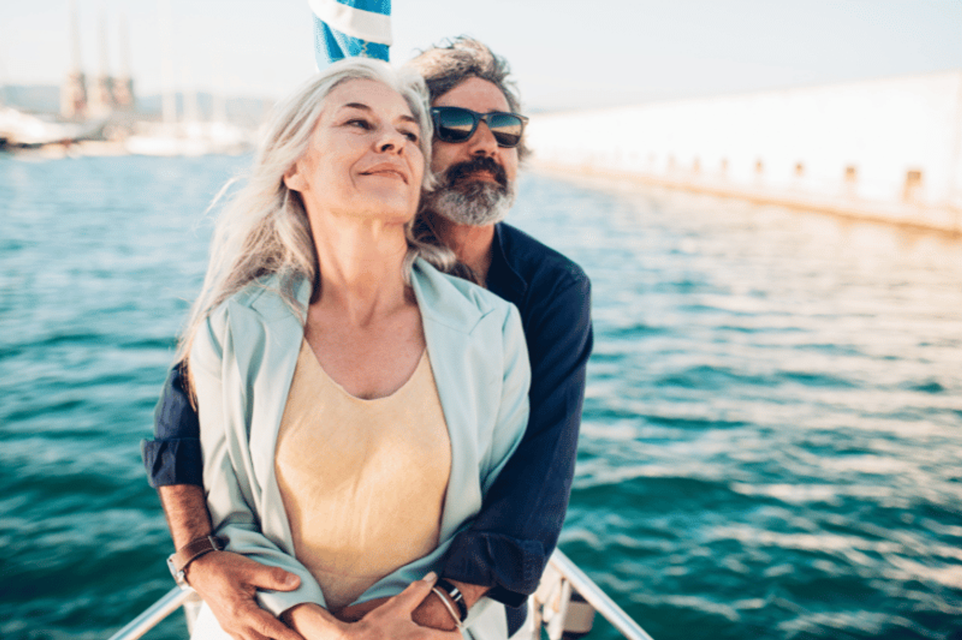 An older couple embracing on the deck of a boat, looking content with the ocean and a distant shoreline in the background.