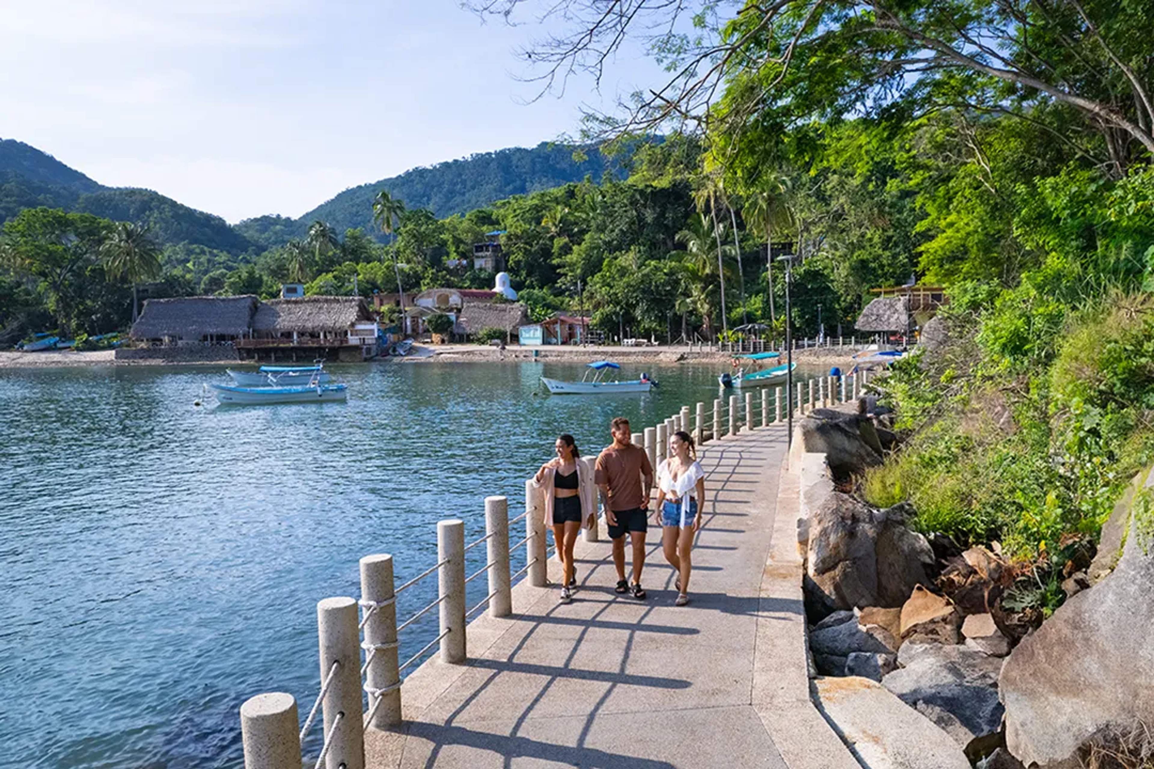 Amigos caminan por un pintoresco malecón con lanchas, palmeras y cerros verdes al fondo.