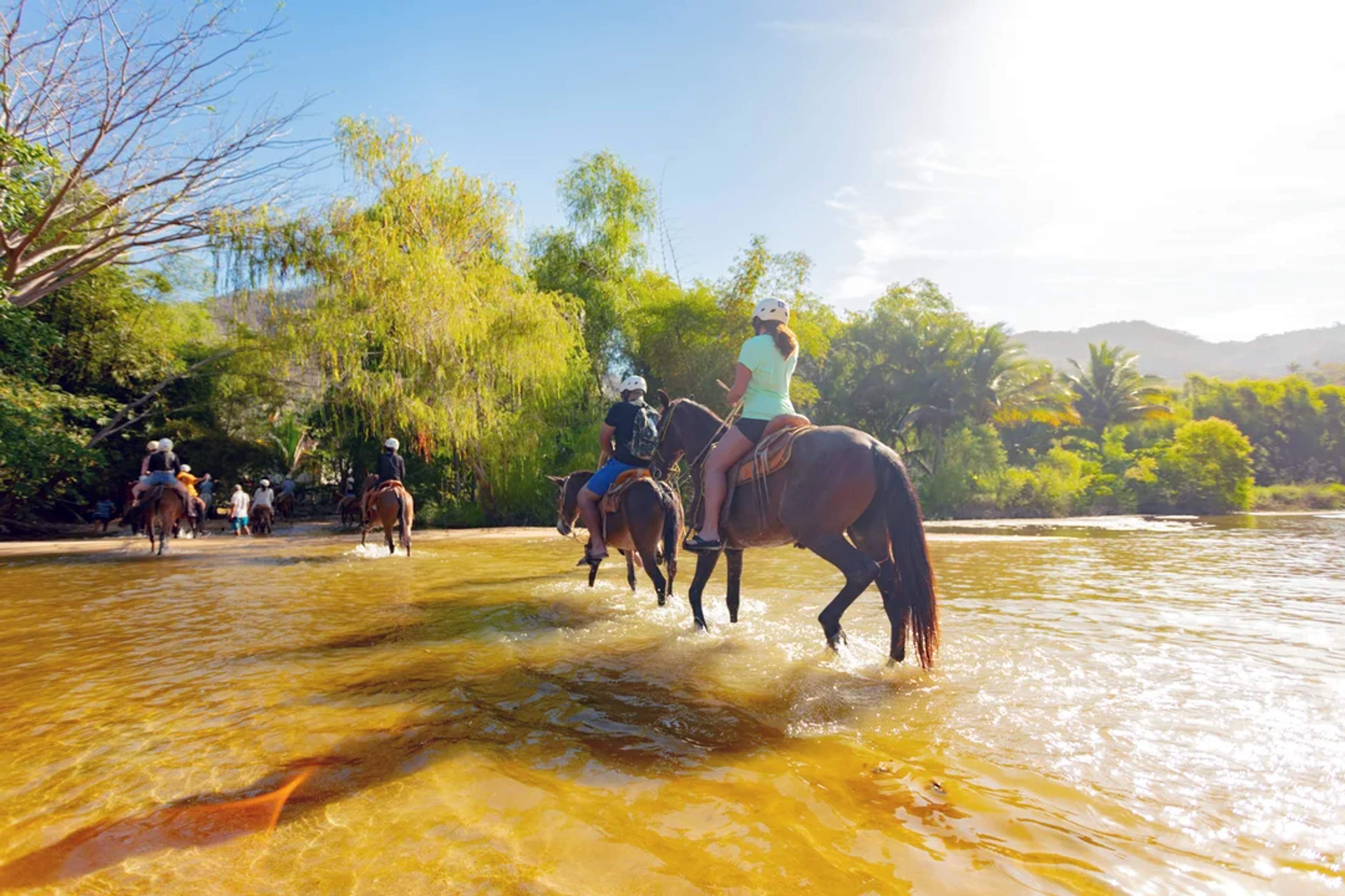 Riders cross a shallow river on horseback, surrounded by lush jungle and golden sunlight.