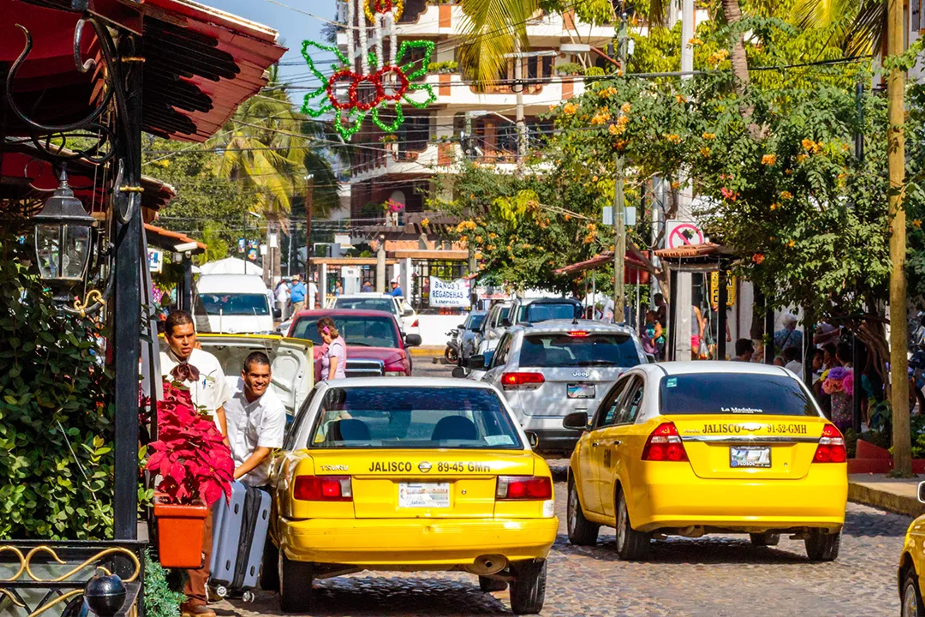 Calle concurrida en Puerto Vallarta con taxis amarillos, adornos festivos y maletas.