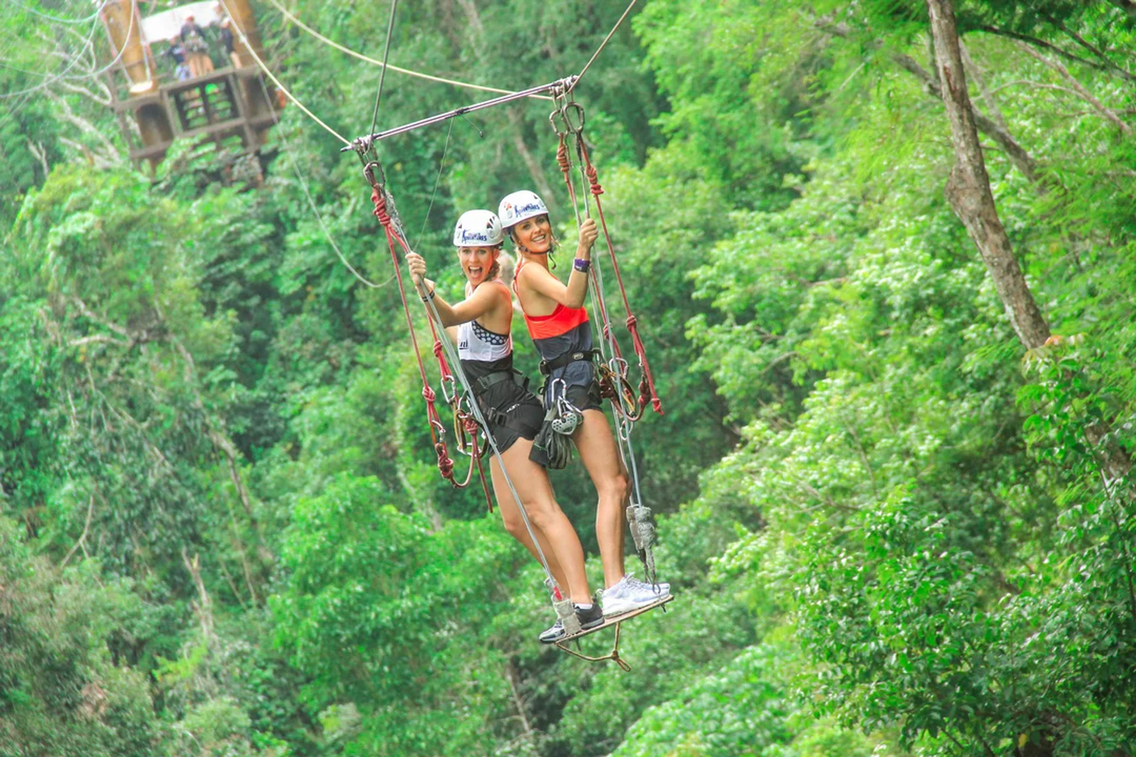 Two women zipline together through lush jungle, smiling and enjoying an adrenaline-filled ride.