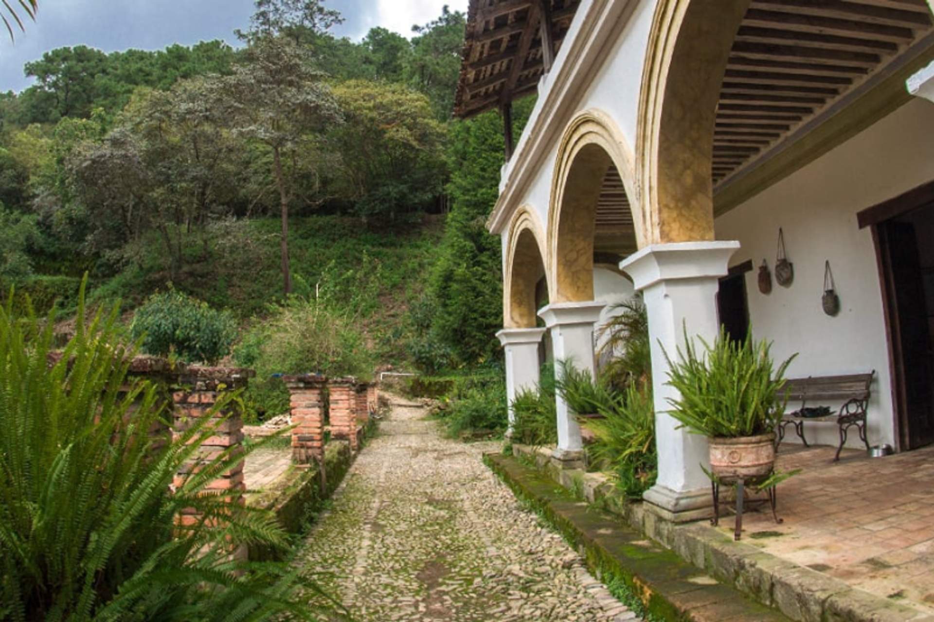 Un camino rústico de adoquines que conduce a un edificio de estilo colonial con pilares arqueados, rodeado de vegetación exuberante y colinas boscosas en el fondo.
