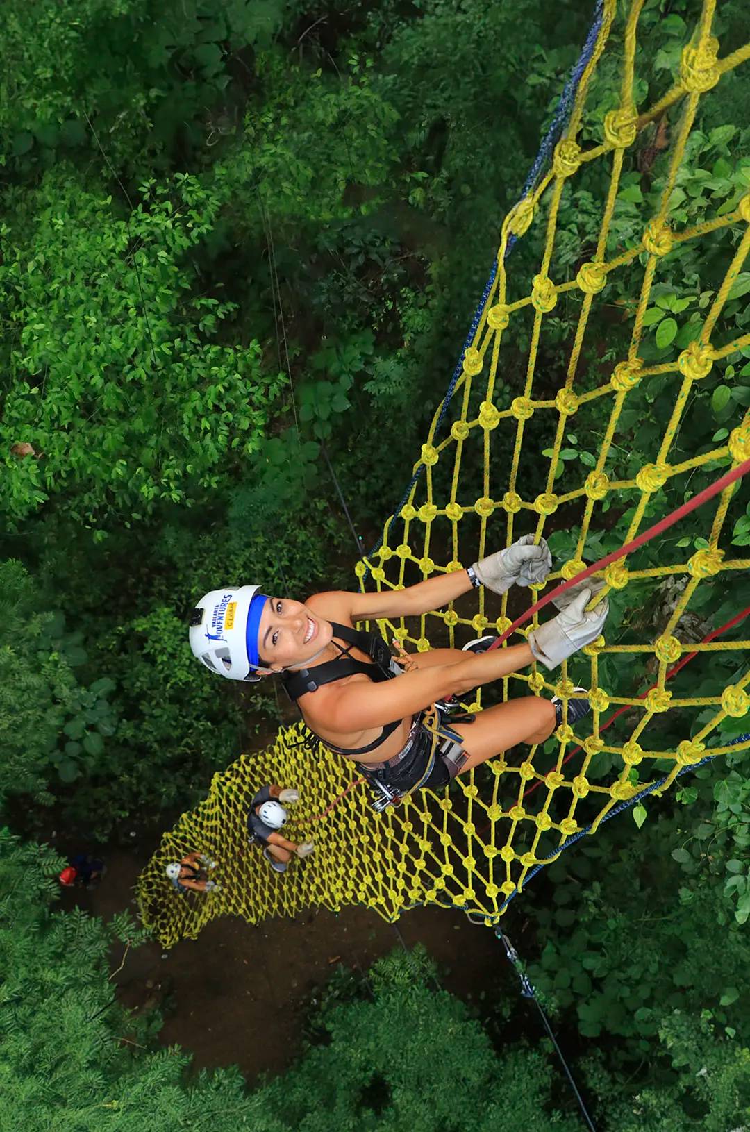 Woman climbs a 65 ft. high yellow rope net amidst lush greenery on the Extreme Adventure tour.