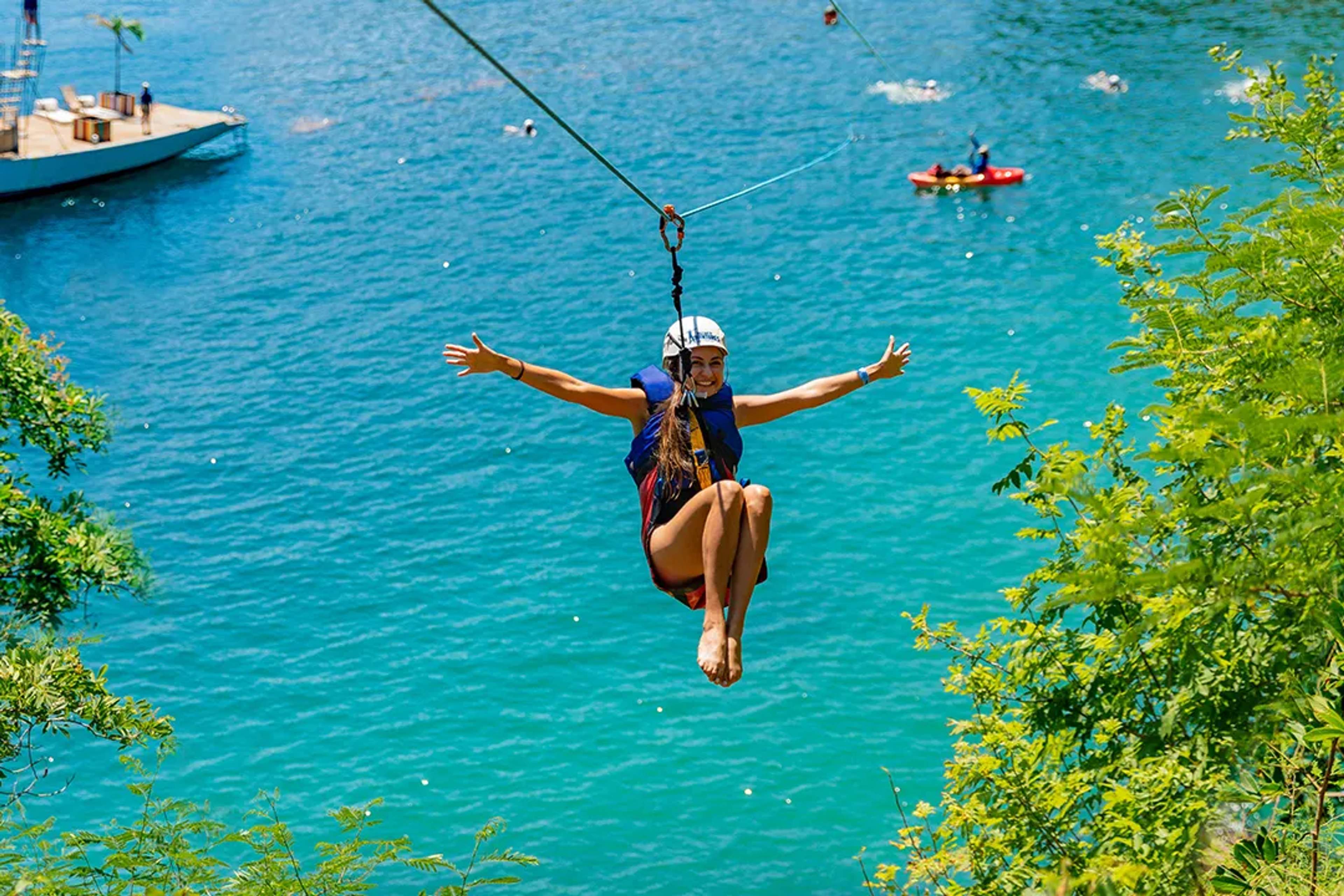 Mujer sonriente en tirolesa sobre agua turquesa, con brazos abiertos, casco y chaleco salvavidas.