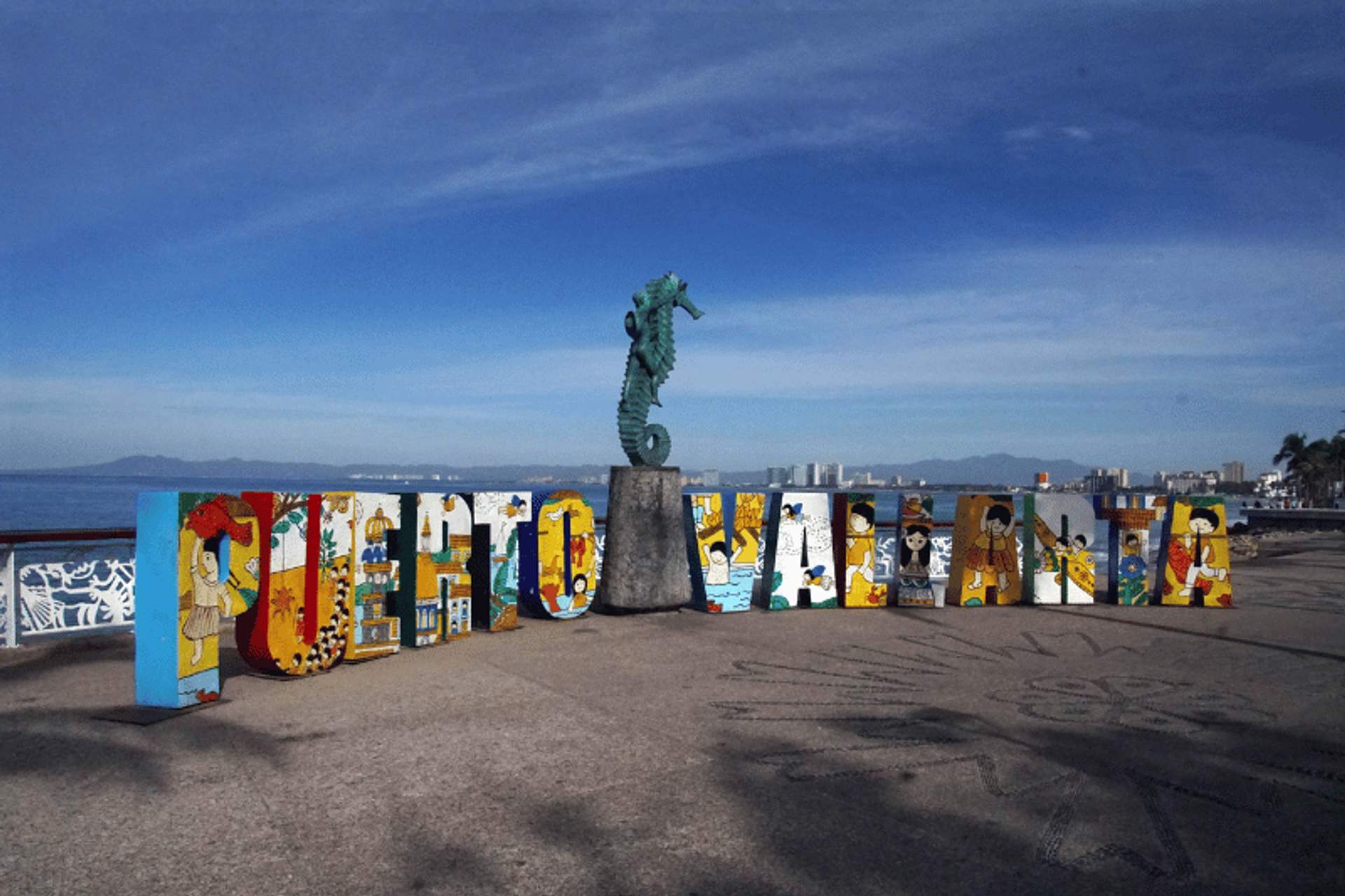 Un letrero colorido que deletrea "Puerto Vallarta" en el malecón, con una estatua de caballito de mar detrás. El letrero está decorado con obras de arte vibrantes que representan varios elementos culturales y naturales de la zona, y el océano y el horizonte de la ciudad son visibles en el fondo.