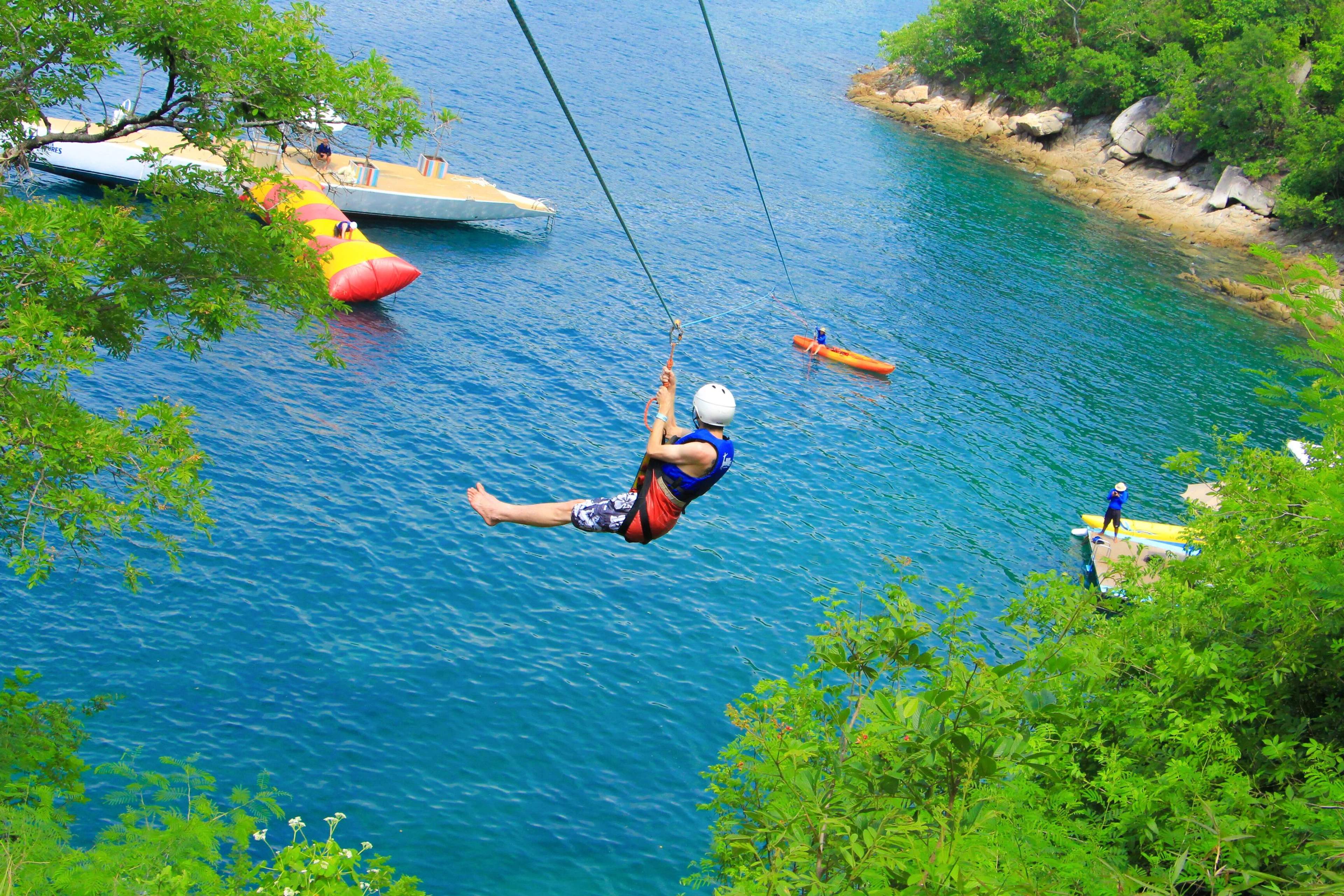 A person ziplining over turquoise water near boats and kayakers, highlighting extreme vacation activities in Puerto Vallarta.