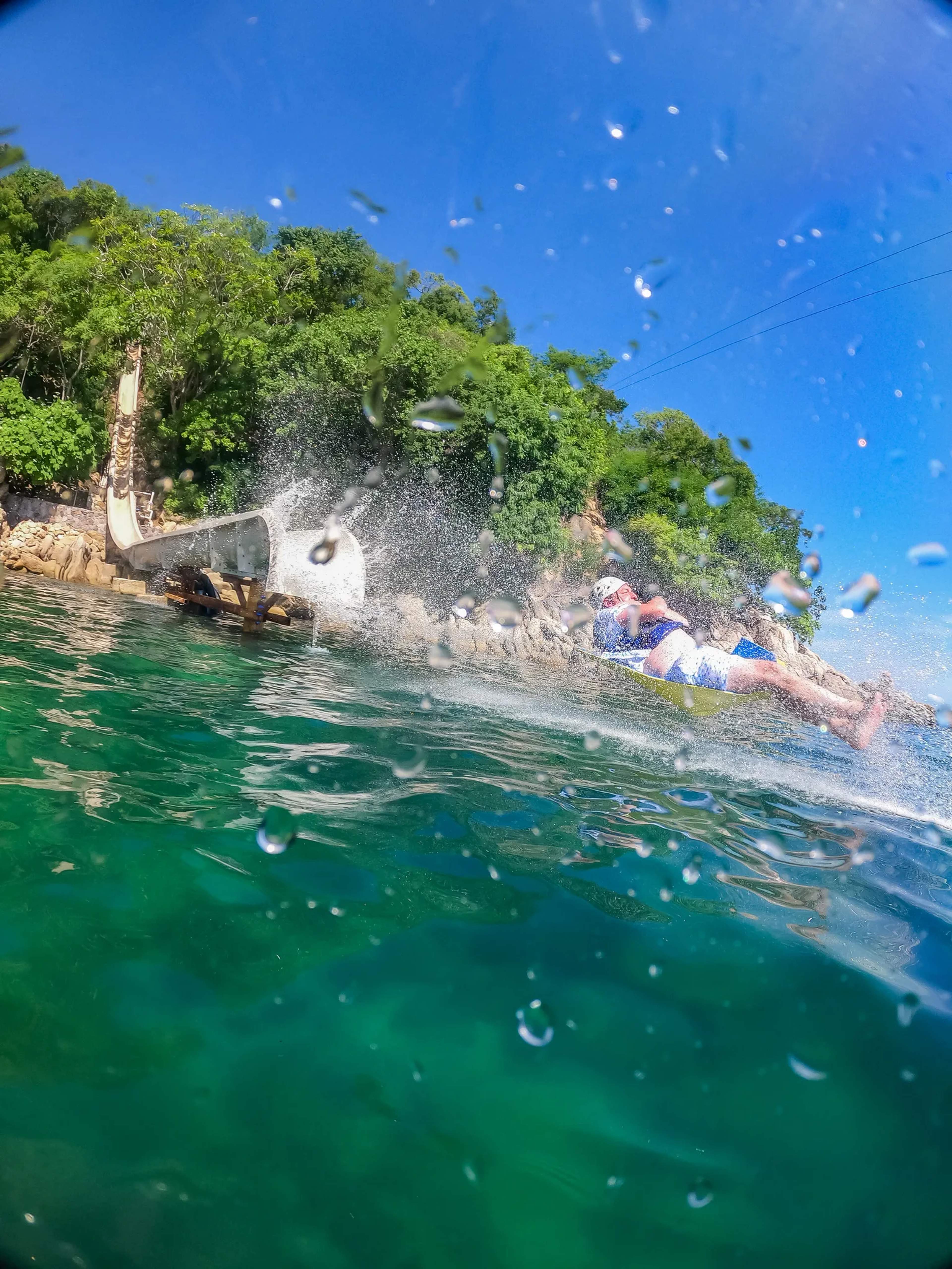 A swimmer enjoys crystal-clear emerald waters beside a lush, vine-covered rocky shoreline under brilliant blue skies.