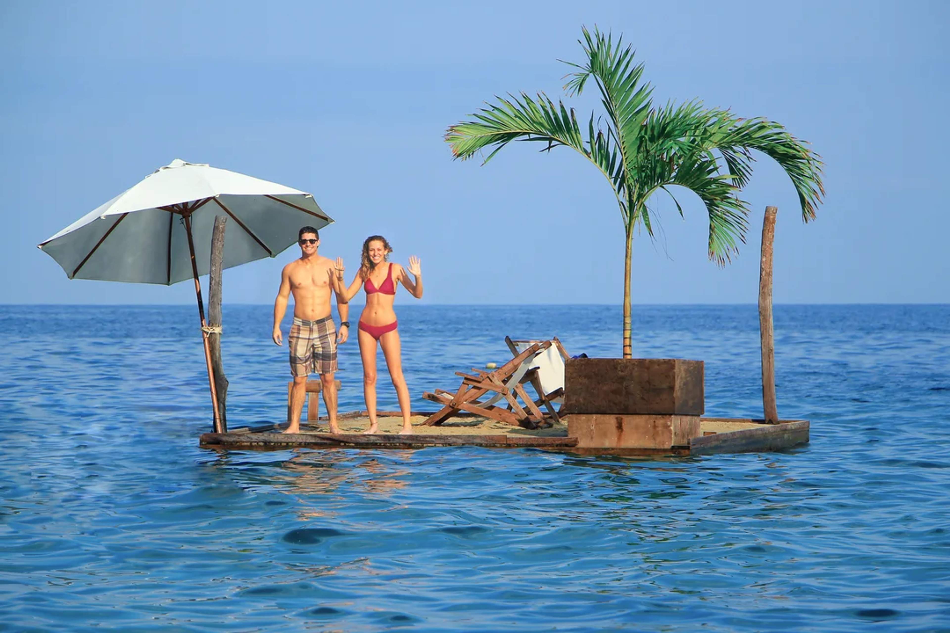 Pareja saluda desde una plataforma flotante con palmera y sombrilla en medio del océano.