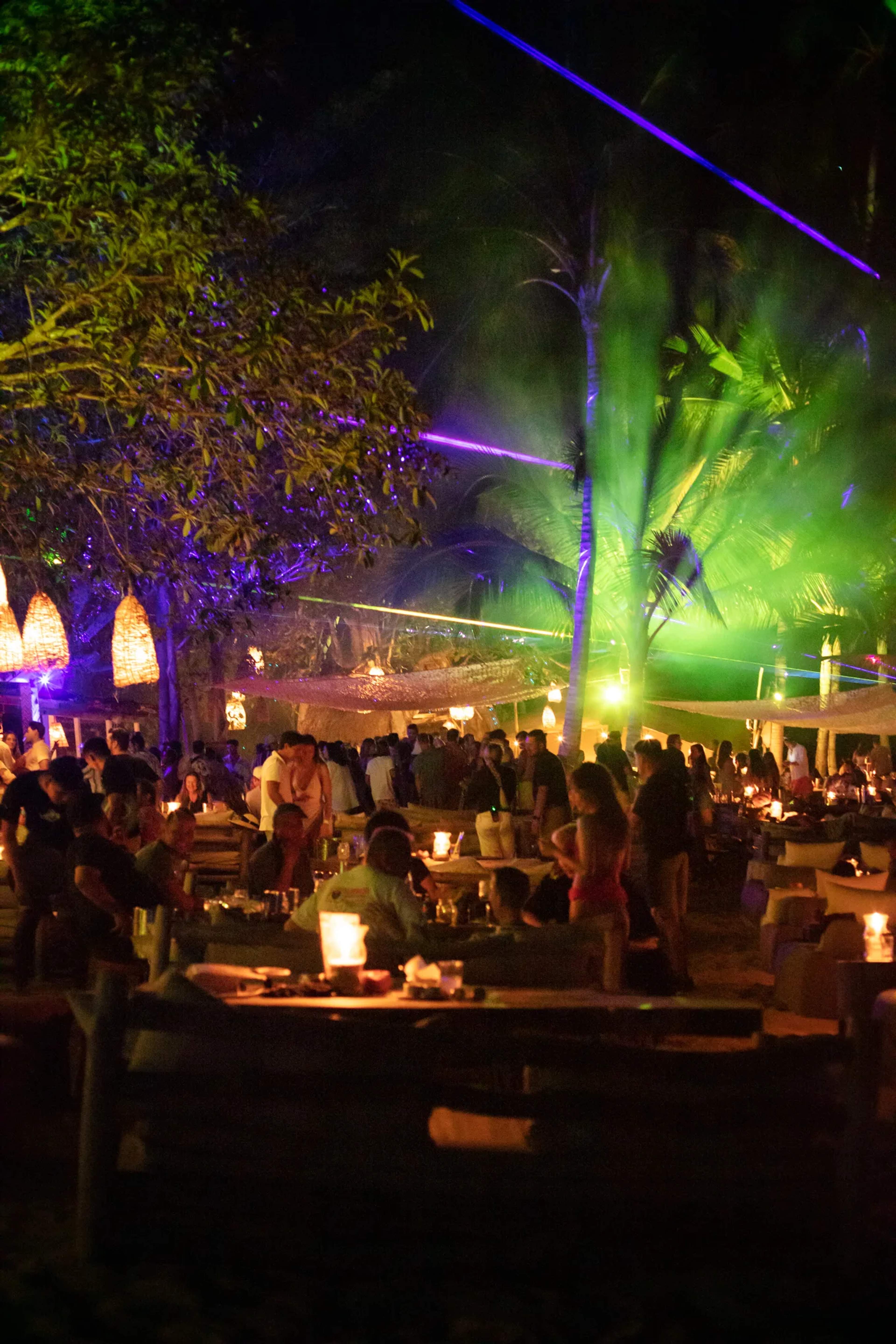 A nighttime beach gathering at Las Caletas lit by purple and green uplighting on palms, with guests mingling at candlelit