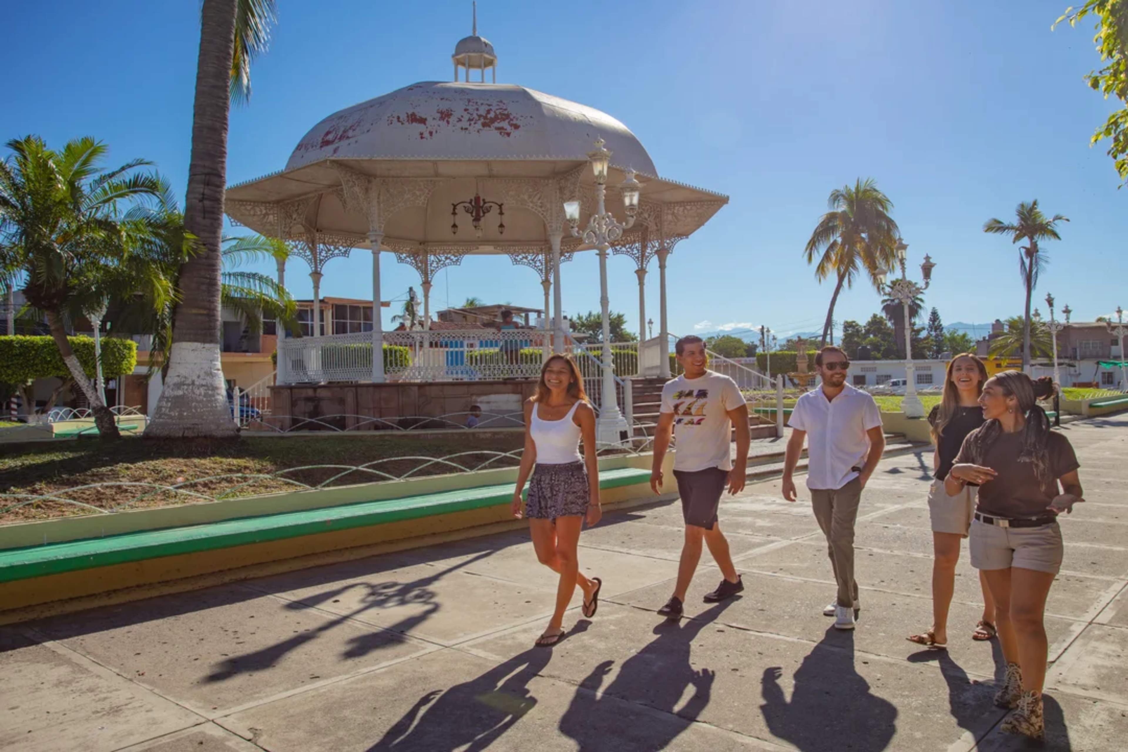 Group of friends strolls through a sunny town plaza with a traditional white gazebo.