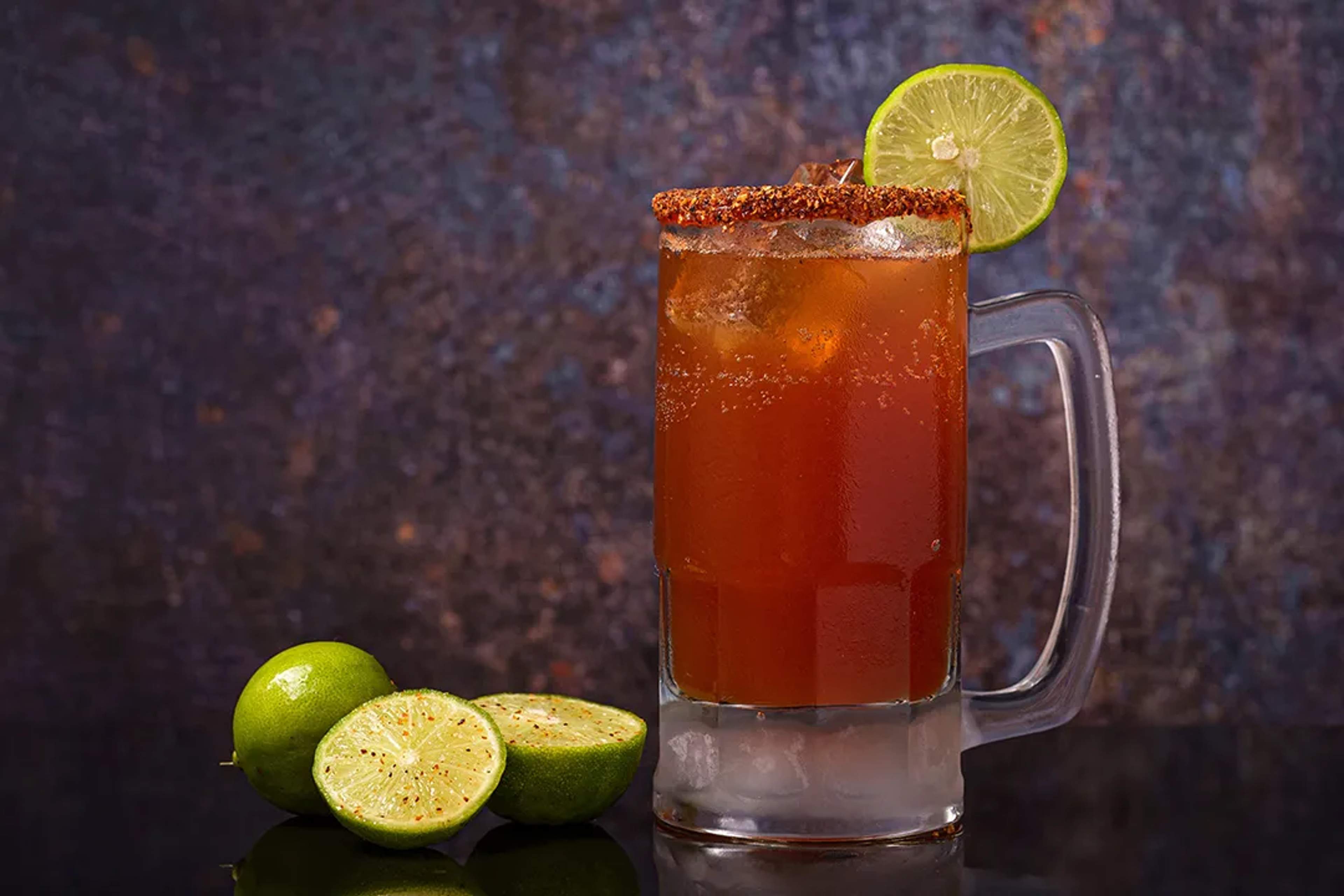 Tarro escarchado de michelada con borde de chile, rodaja de limón y limones frescos al lado.