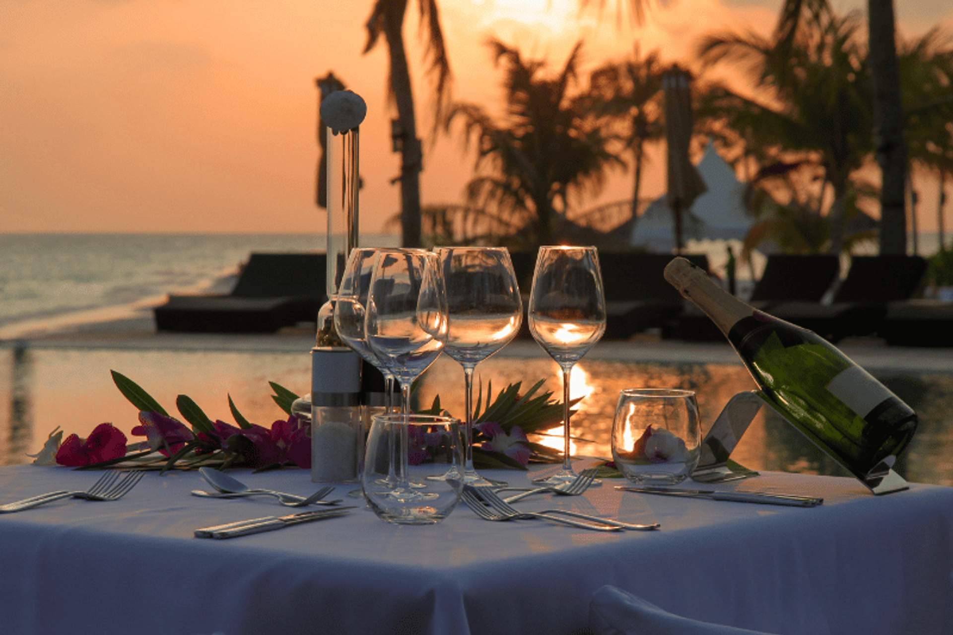 A romantic dinner setup by the beach at sunset, featuring wine glasses, a bottle of champagne, and tropical flowers.