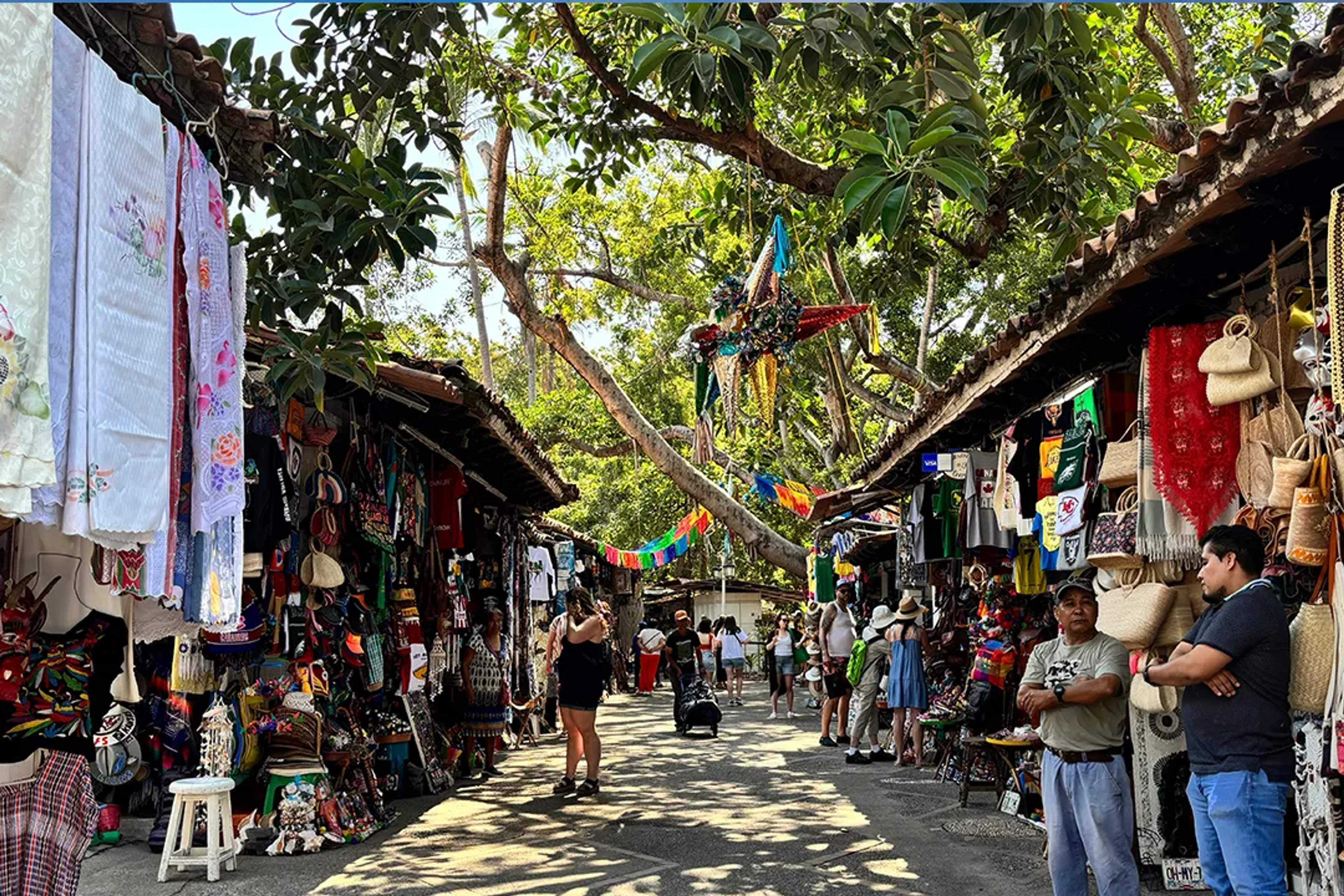 Mercado mexicano colorido con artesanías, ropa y decoraciones bajo árboles frondosos.