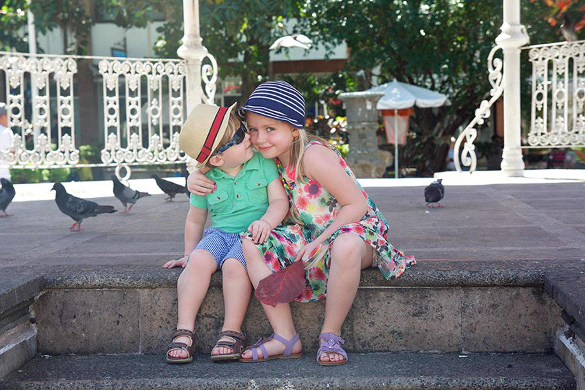 Dos niños con ropa de verano y sombreros sentados en escalones de piedra en el centro de Puerto Vallarta, con palomas al fondo, compartiendo un momento tierno.