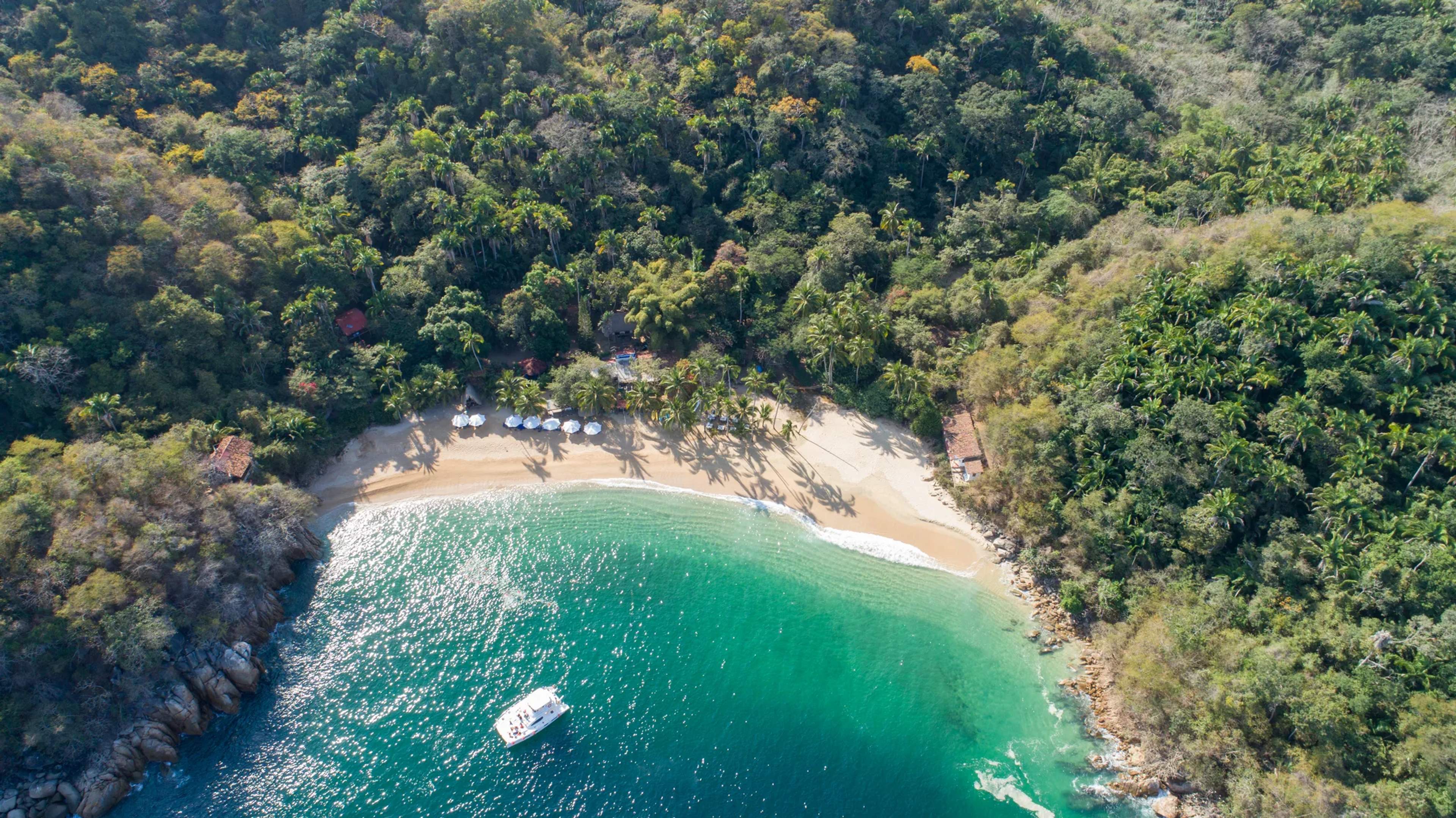 Aerial view of a secluded tropical beach cove with turquoise waters, white sand, dense jungle, and a small white boat