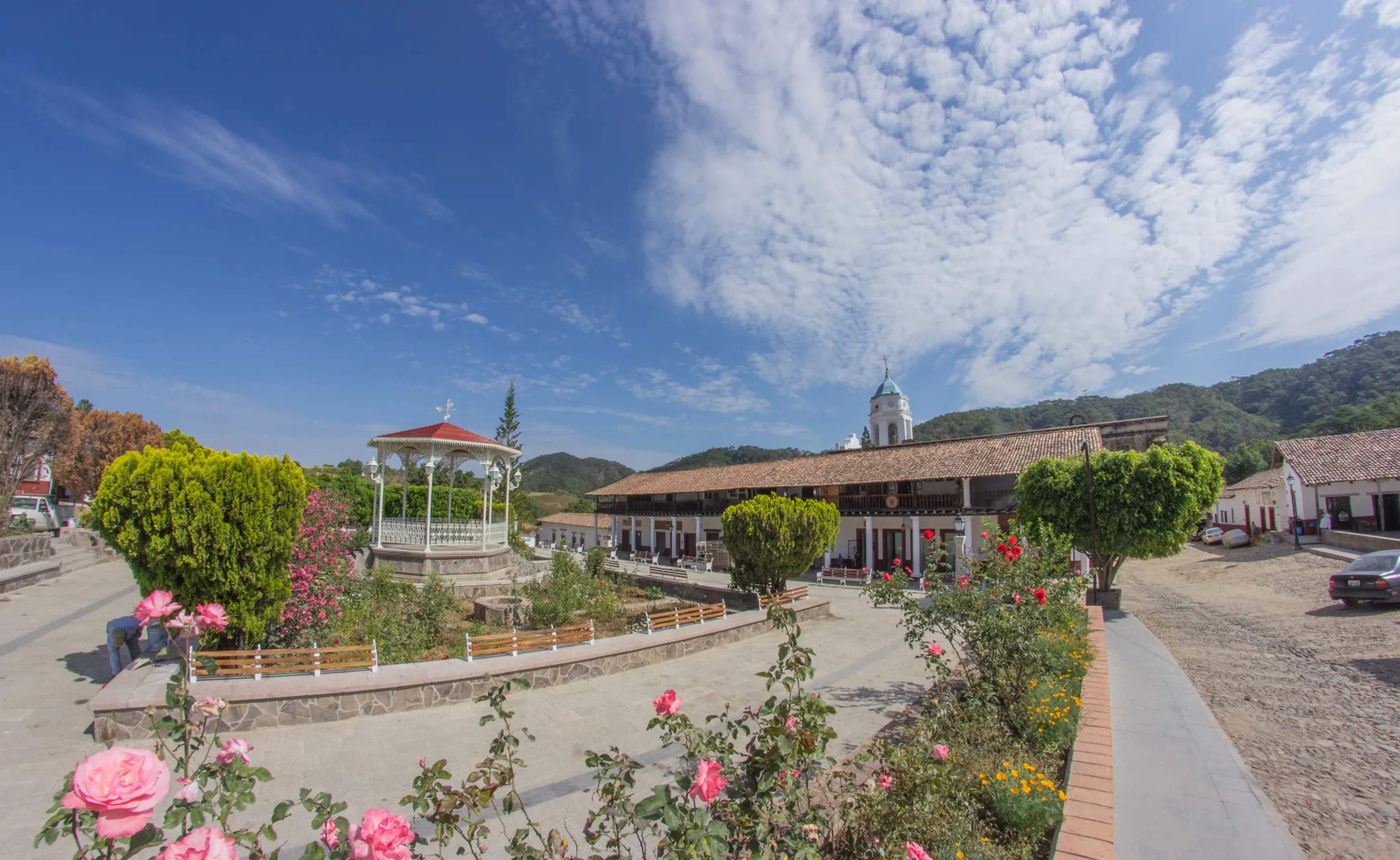 Colonial Mexican village square with a white gazebo, bougainvillea, and a blue-domed church set against mountains and clear