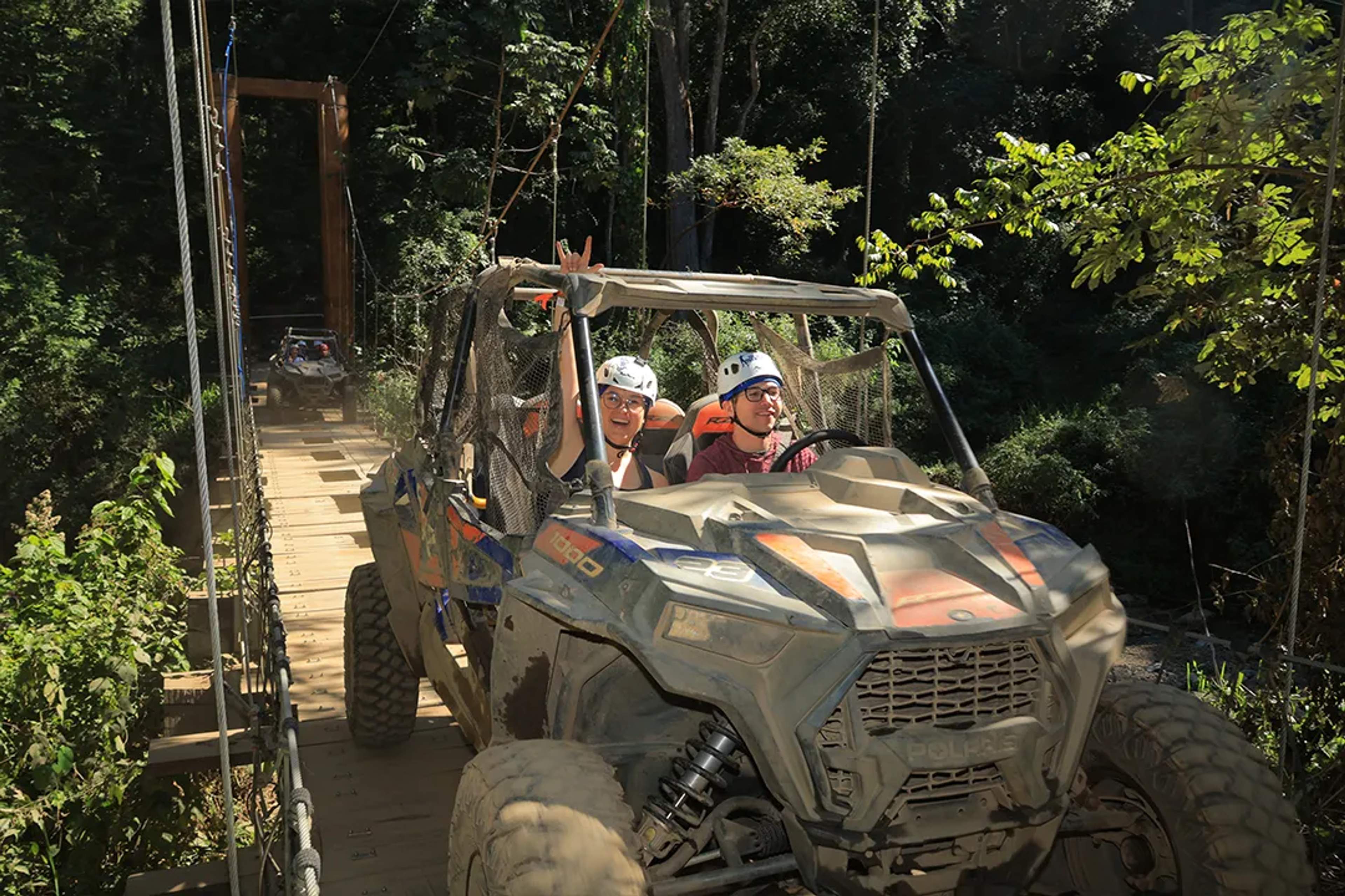 Adventurers cross a jungle suspension bridge in a Polaris RZR, smiling through the off-road thrill at Extreme Adventure.