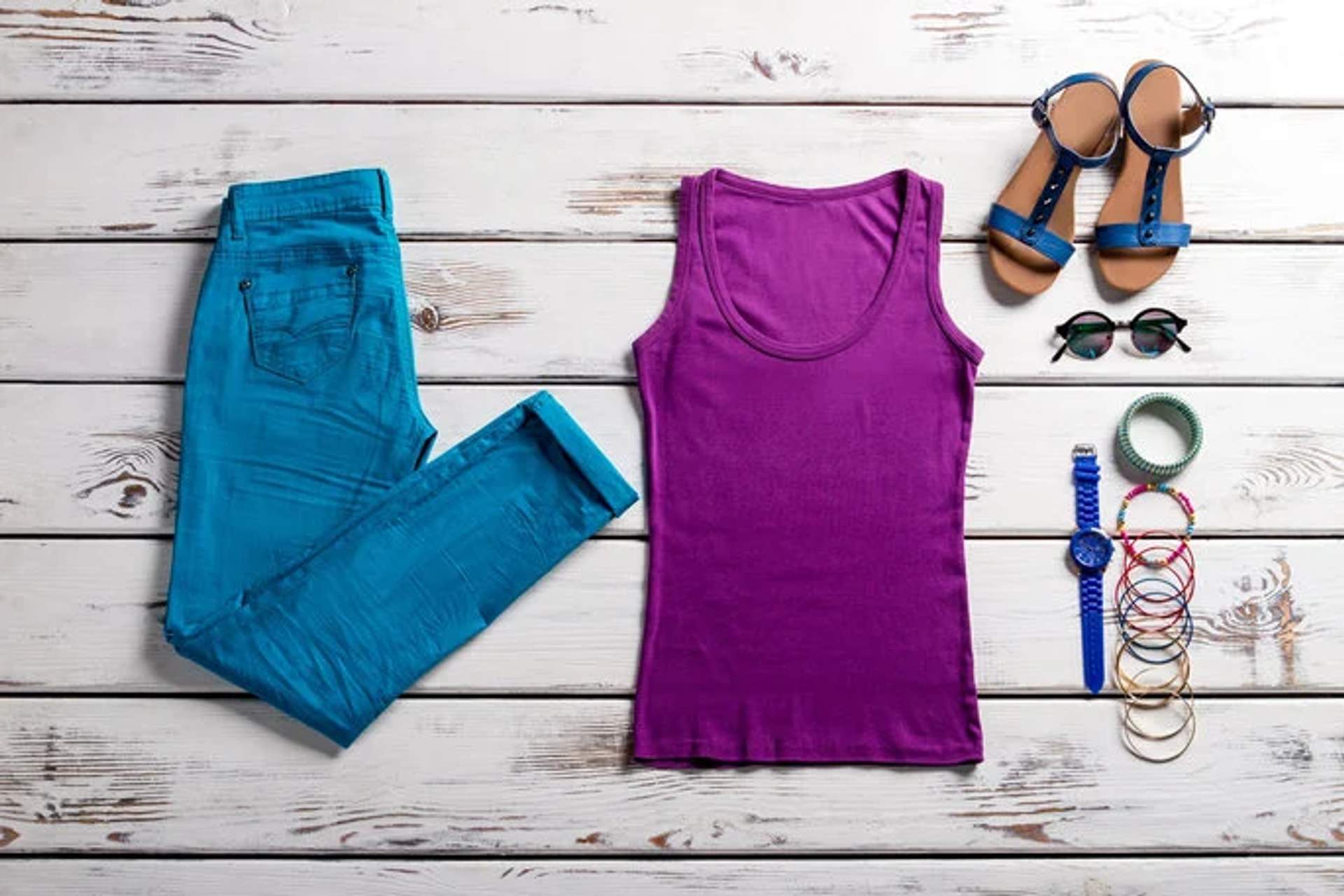 Casual outfit laid out for packing, including turquoise jeans, purple tank top, sandals, sunglasses, and accessories.