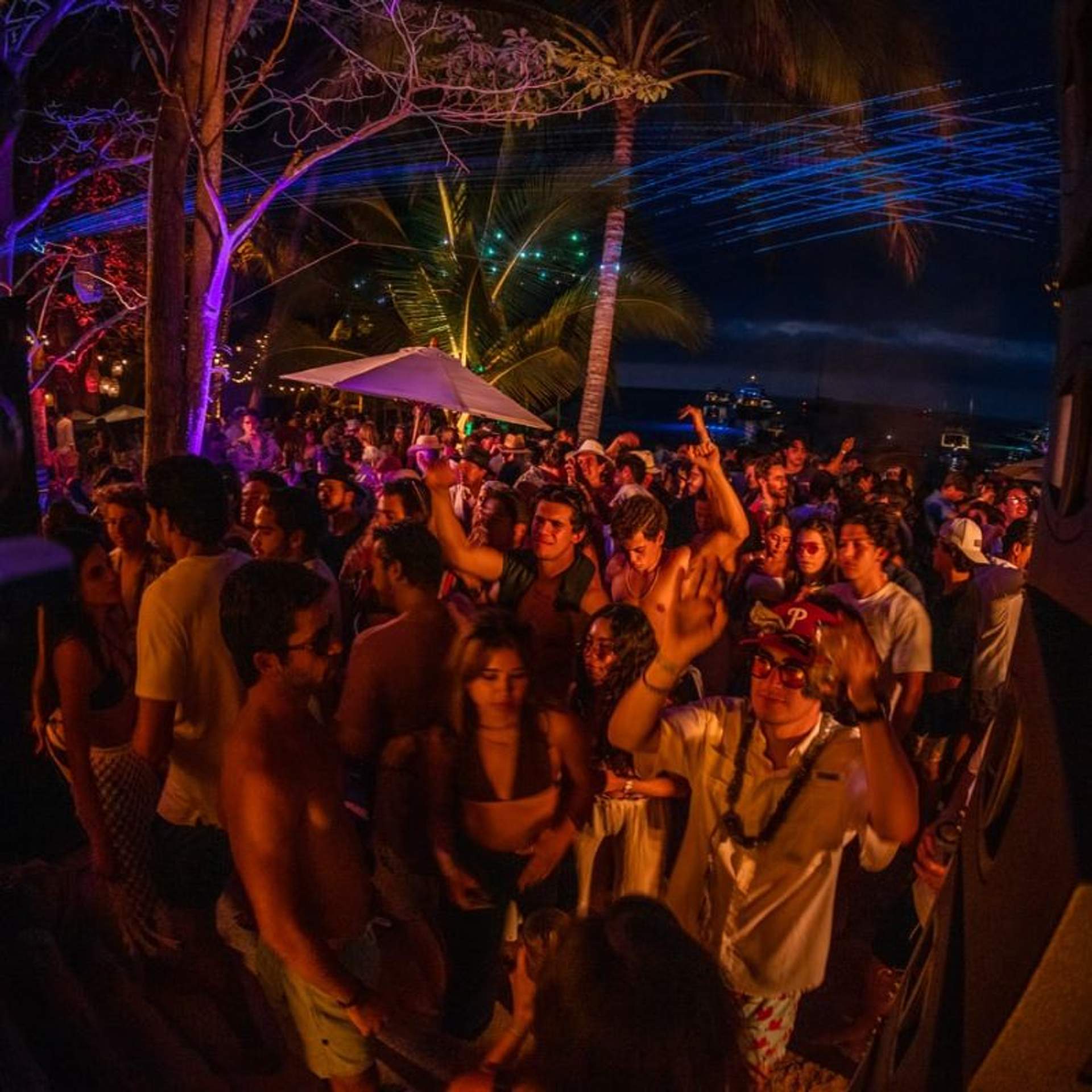 A lively beach party at night with a large crowd dancing under colorful lights and palm trees in the background.
