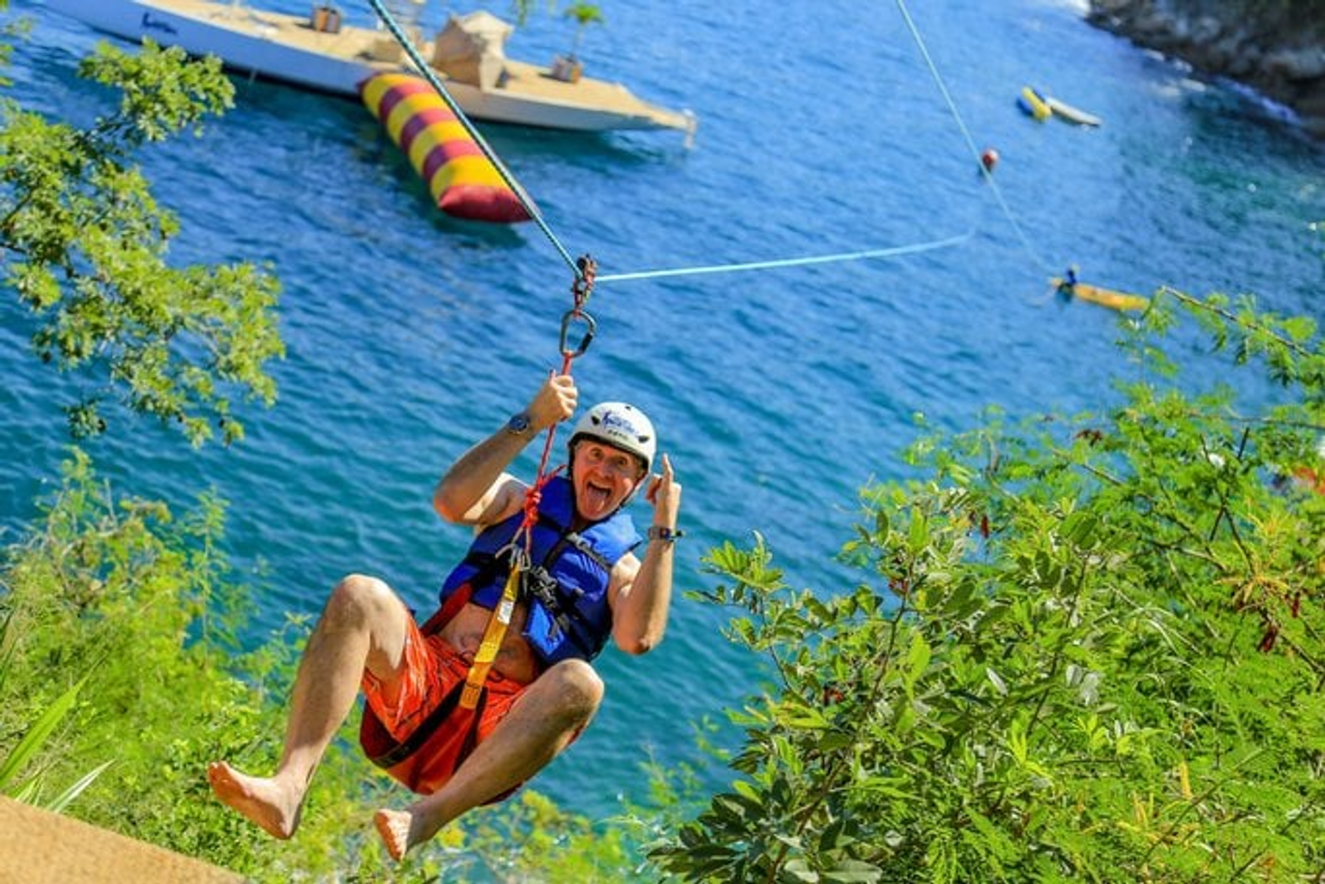 Un hombre haciendo tirolesa sobre el agua, sonriendo y haciendo un gesto juguetón con la mano. Lleva un casco y un chaleco salvavidas, con una gran estructura inflable visible en el agua abajo.