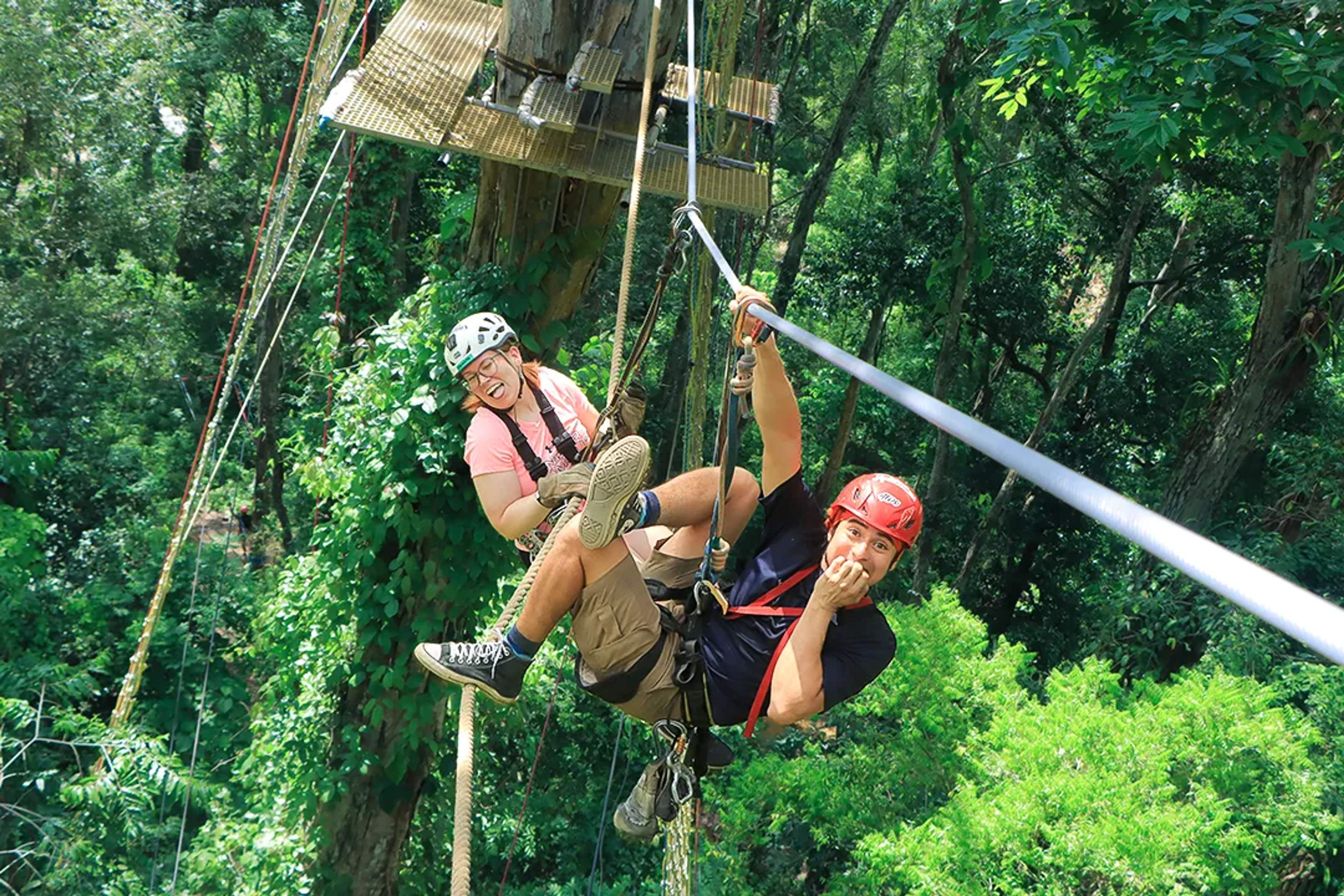 Two adventurers dangle mid-zipline, laughing and posing playfully above the lush jungle canopy at Extreme Adventure.