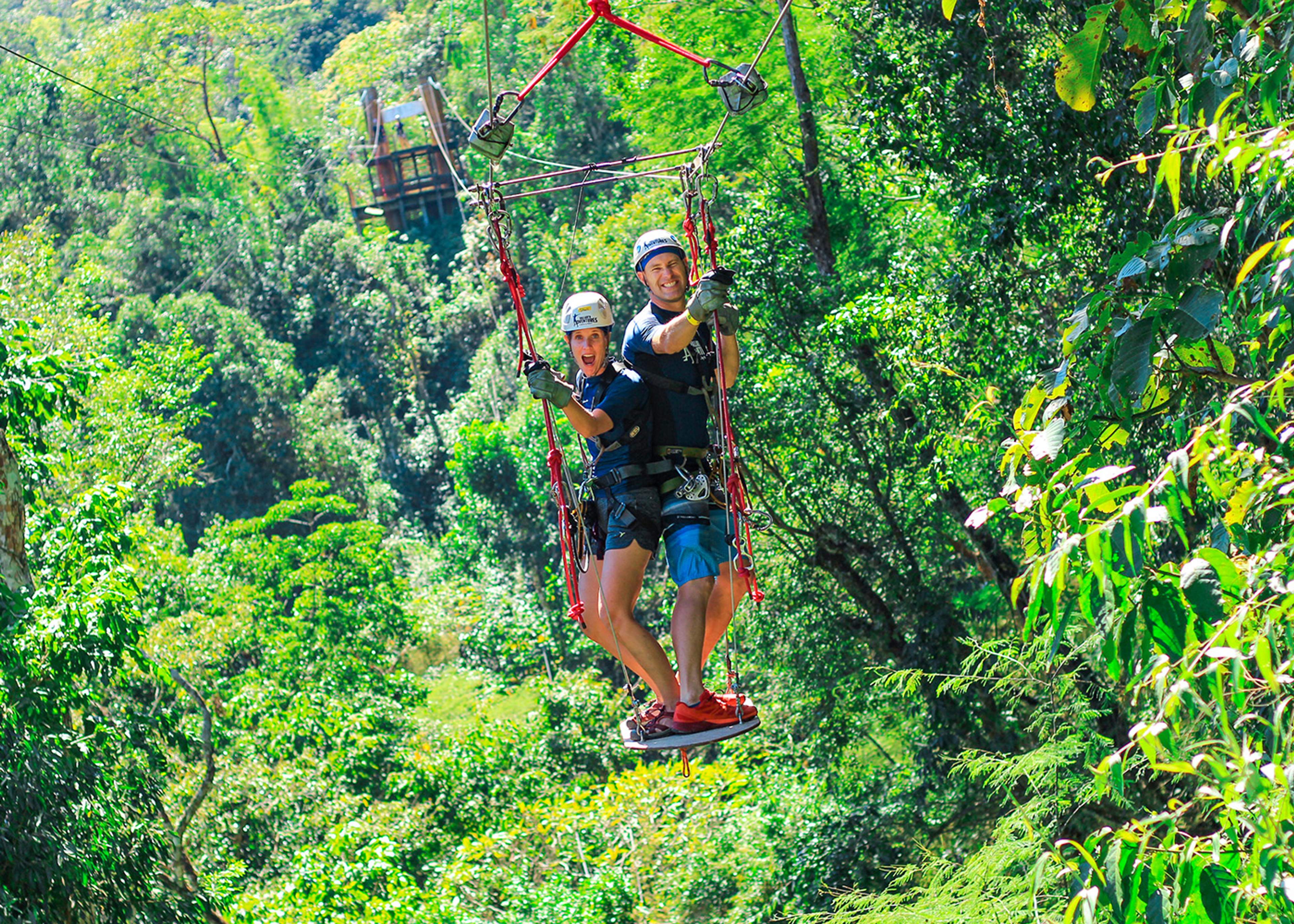A smiling couple wearing helmets and harnesses enjoys ziplining together through a lush forest, standing on a platform suspended by ropes.