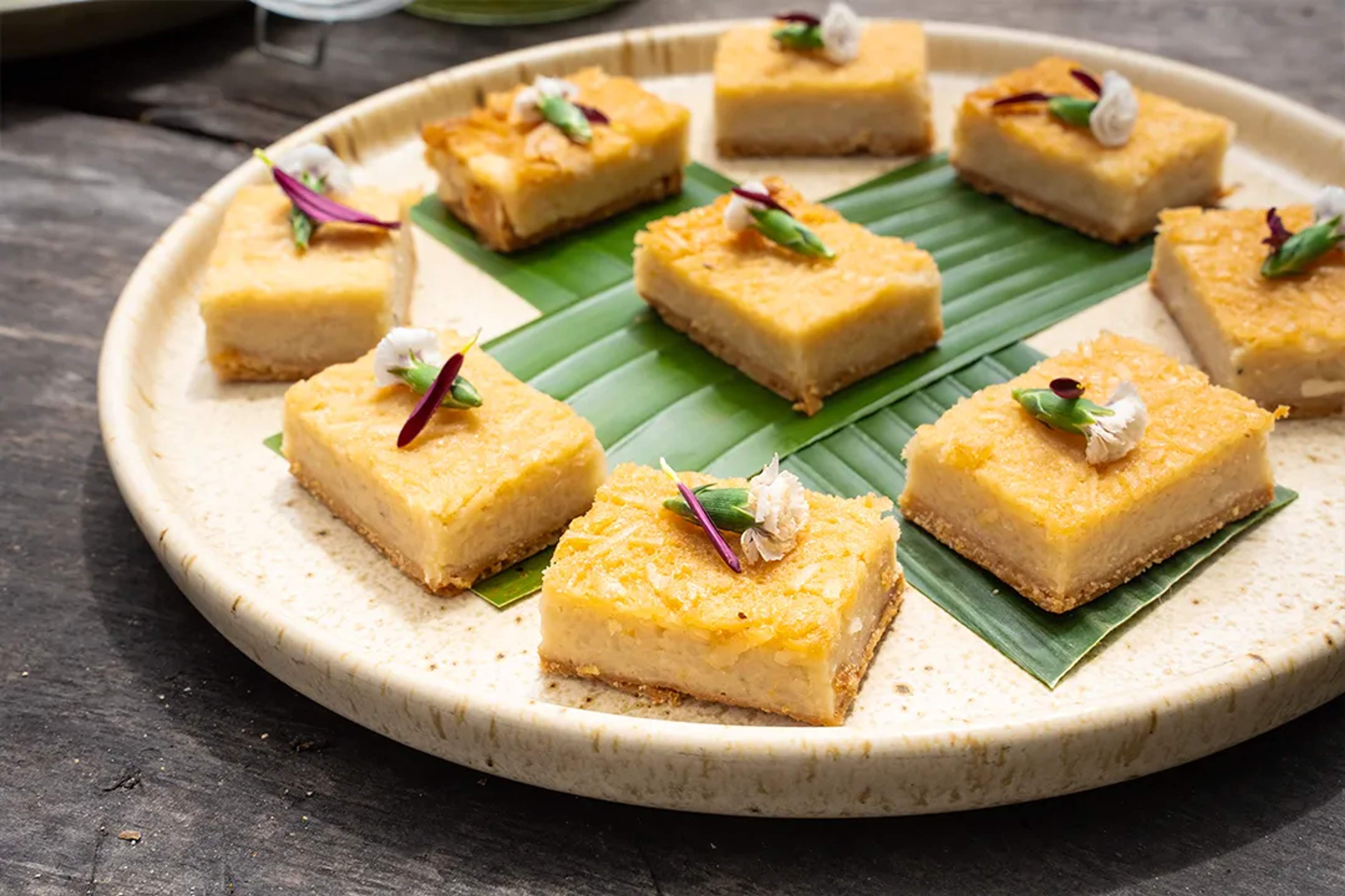 Cuadritos de postre de coco decorados con flores, servidos sobre hojas de plátano.