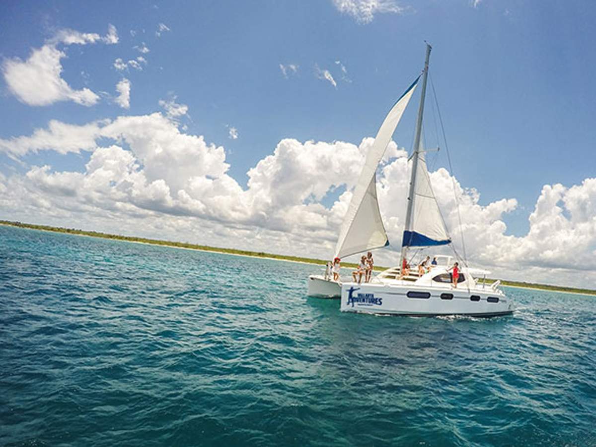 A sailboat with people on deck cruising on clear blue waters under a bright sky with scattered clouds.