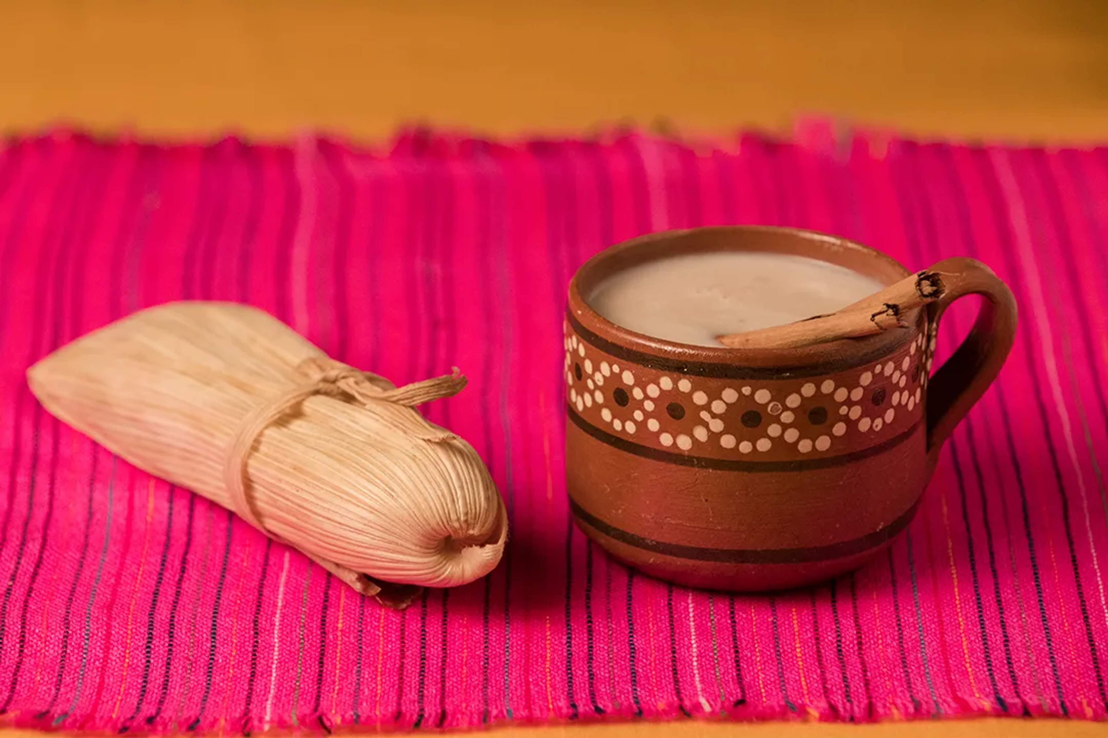 Jarro de barro con champurrado y un tamal envuelto en hoja de maíz sobre textil rosa.