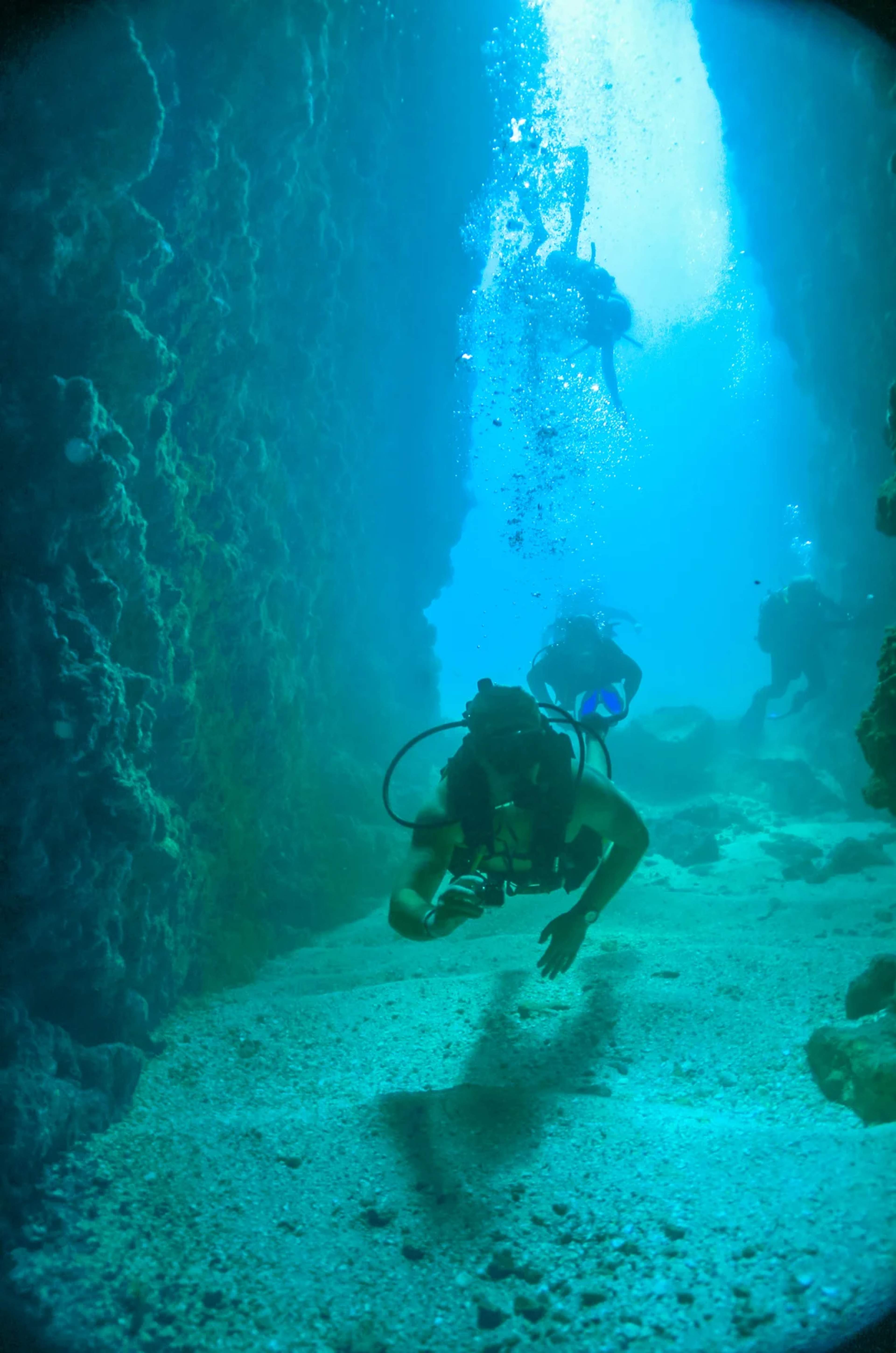 Divers explore an underwater canyon surrounded by rocky walls and glowing turquoise light.