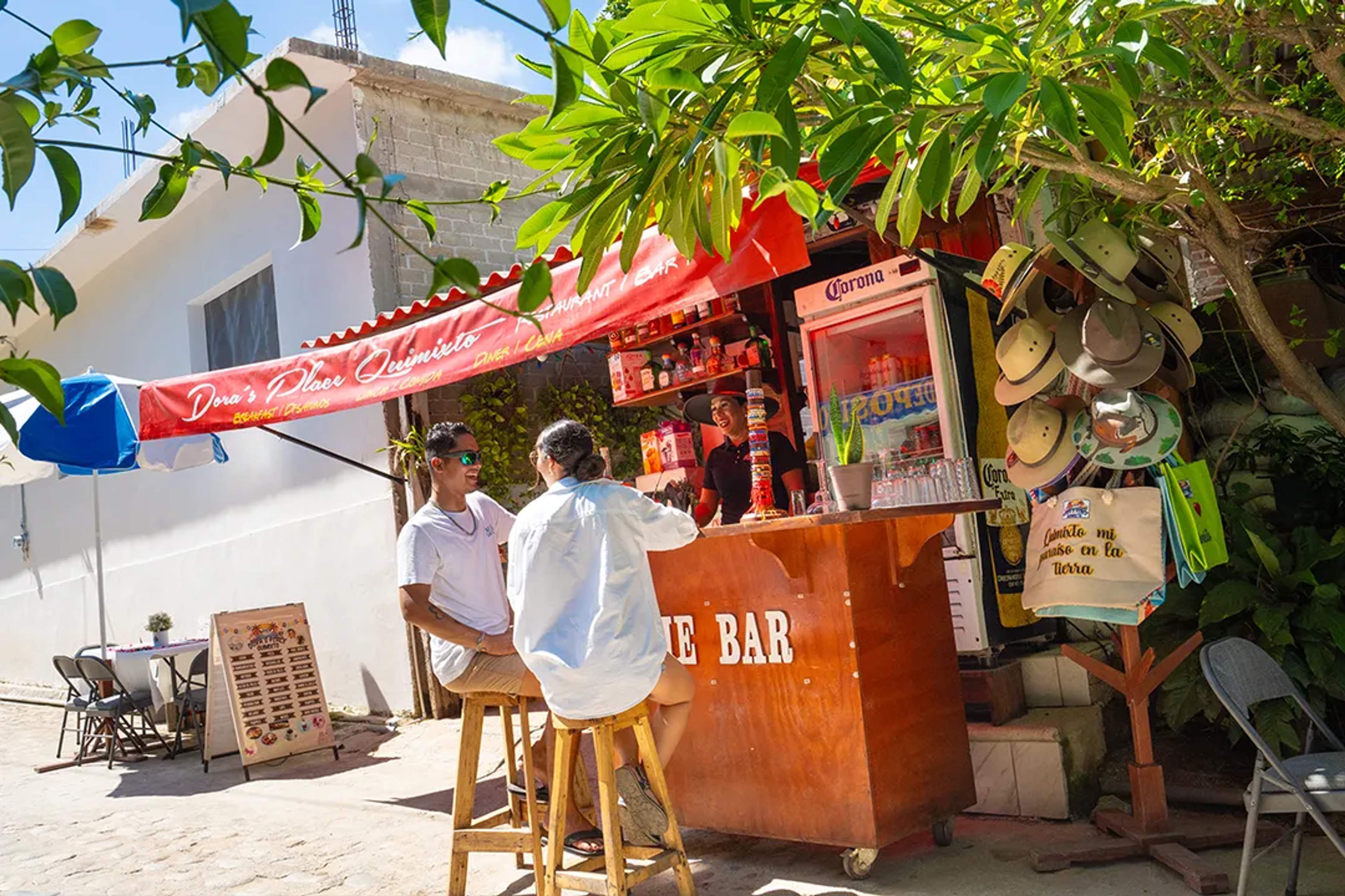 Pareja disfruta bebidas en un colorido bar callejero en Quimixto, bajo sombra y ambiente local.