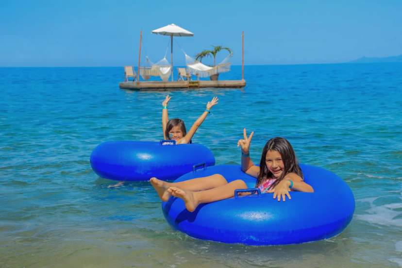 Two happy girls float in the sea with blue tubes near a tropical wooden raft with hammocks.