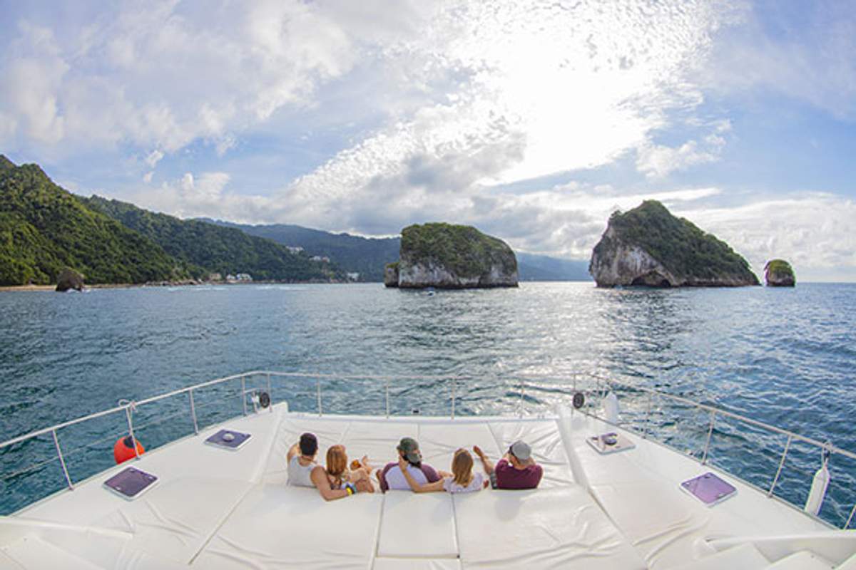People relax on a boat's deck with a view of Los Arcos rock formations in Puerto Vallarta under a partly cloudy sky.