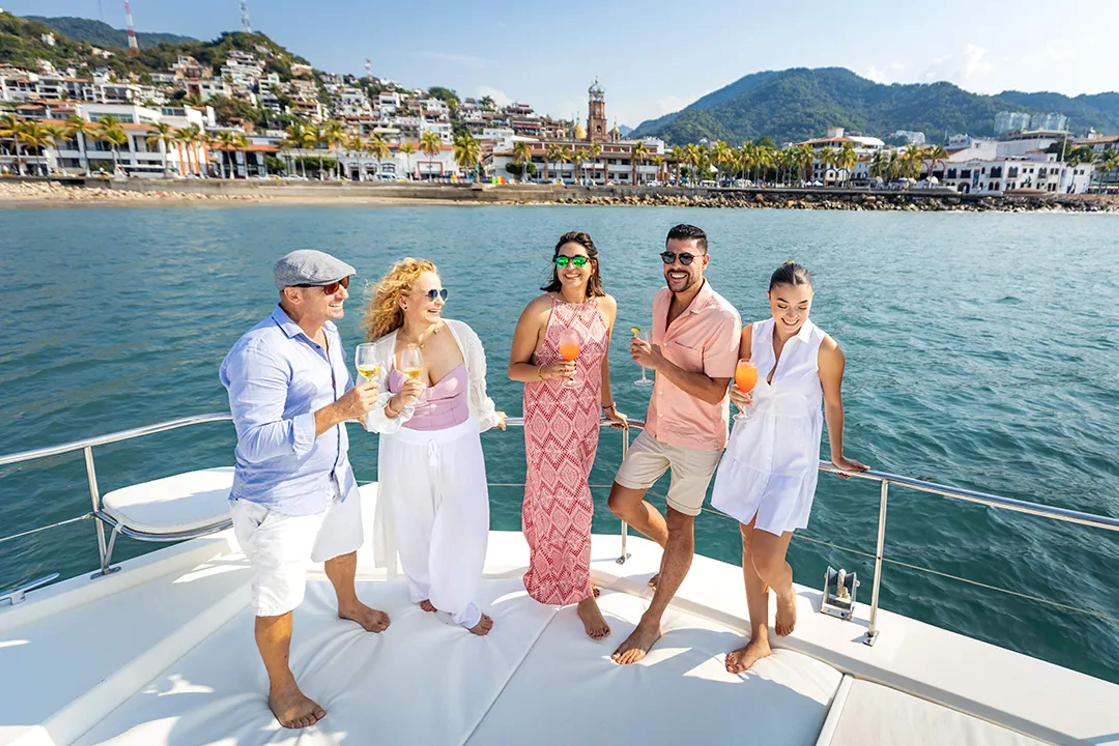 Six guests toast cocktails on a catamaran deck with Sierra Madre mountains and Puerto Vallarta coastline at sunset.