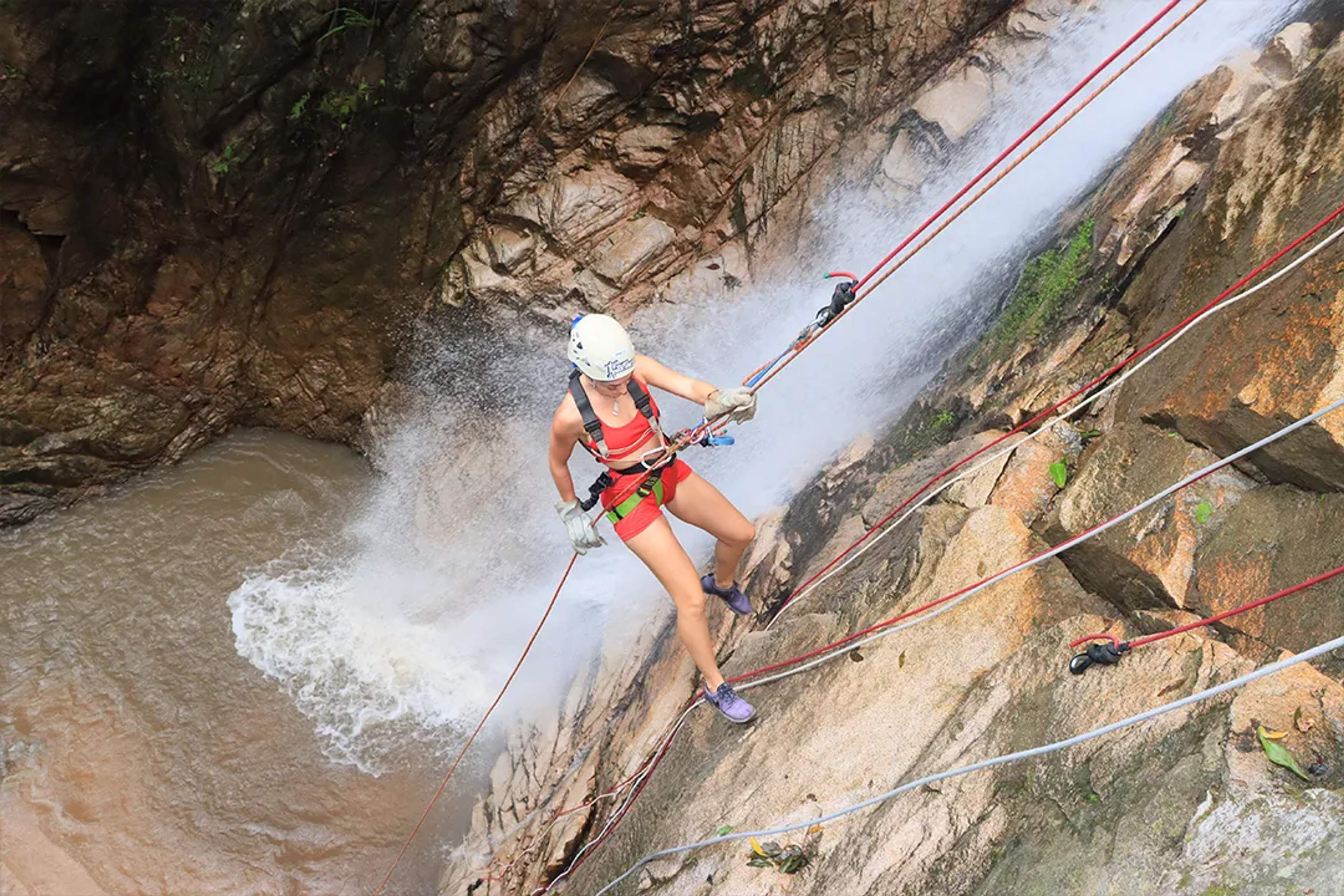 Woman rappelling down a waterfall on a rocky cliffside, wearing safety gear and a helmet.