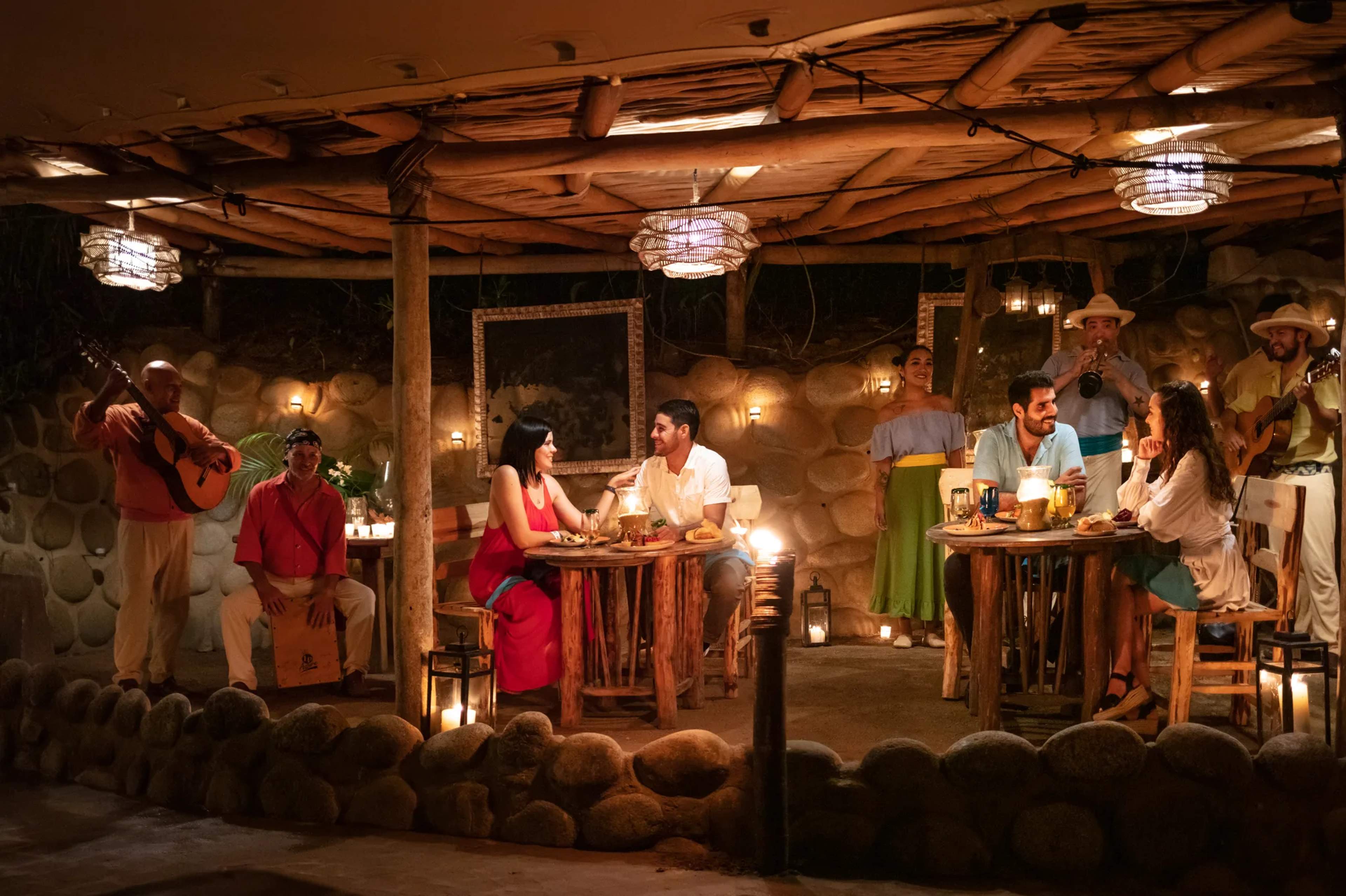 Evening outdoor fiesta with guests enjoying food, drinks, and live music under warm string lights and wooden beams.